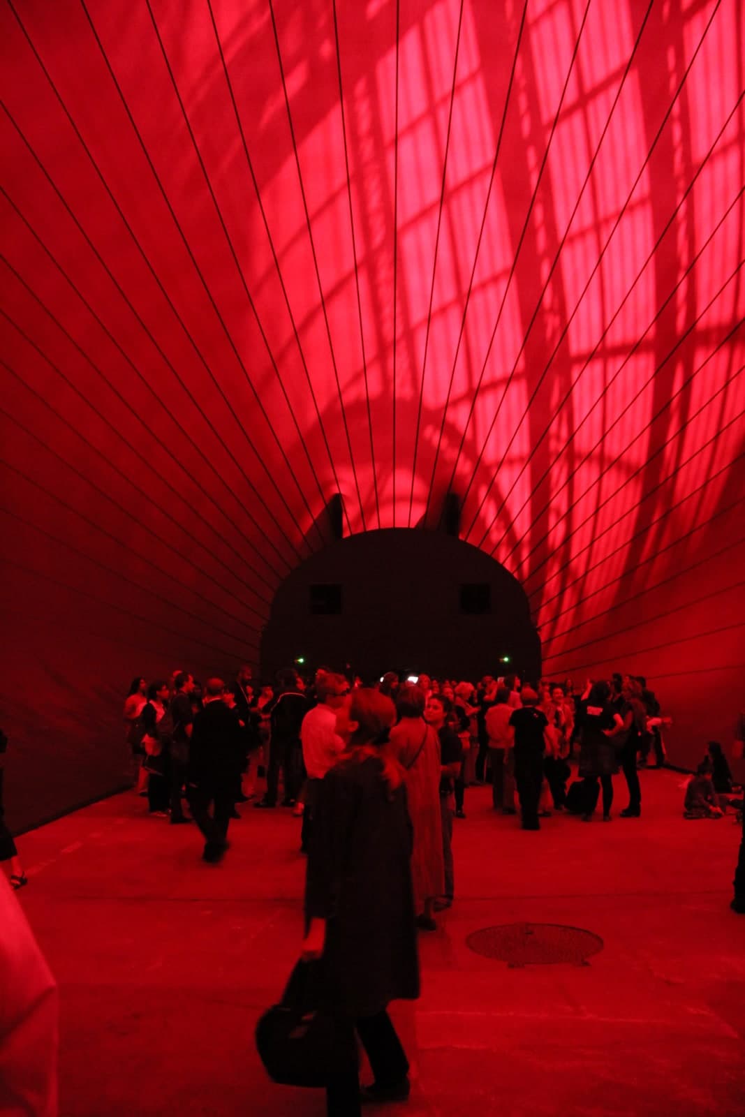 The inside of a vast inflated membrane, lit deep red by daylight coming through it. The wall curves up and over the camera on both sides and meets in a high vault, divided by dark seam lines that run from the floor to the top of the frame. The girders and glazing bars of a glass roof show through the membrane as blurred shadows. At the far end a rounded dark mass rises from the floor with two small pointed projections on top and two rectangular openings in its face. Several dozen visitors stand on the pale concrete floor beneath it, their clothes and skin washed red, with a round metal drain cover in the floor at the right