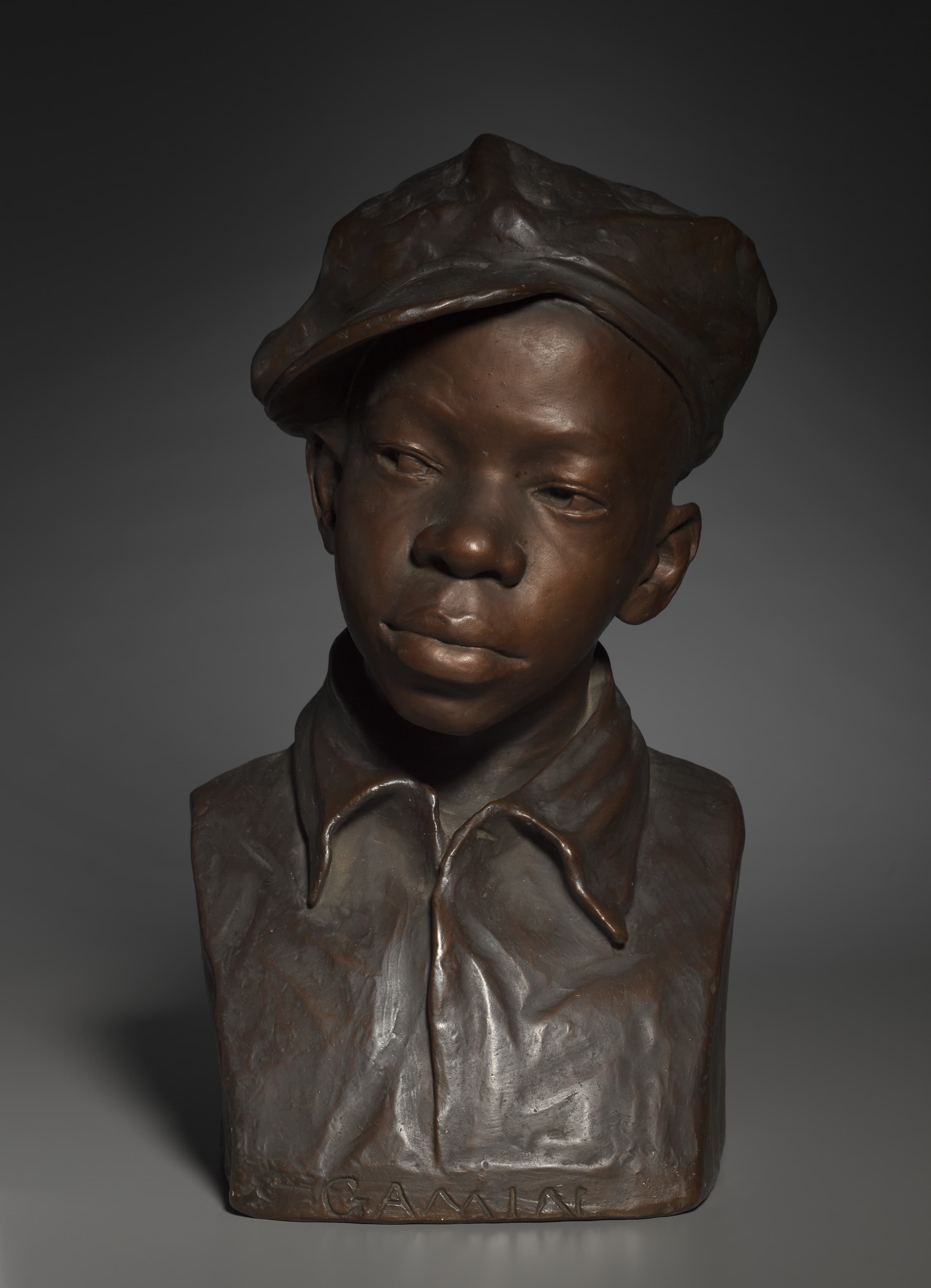 A bust of a boy in a soft flat cap and an open-collared shirt, modelled from the chest up and painted a warm coppery brown. The cap's crown is crumpled and pushed back off the forehead; the brim lifts clear of the brow. His head is turned slightly to his left and tipped down, the eyes half-lidded and the mouth closed. Two long pointed collar tips fall across the chest, and the word GAMIN is cut in block capitals across the bottom front of the base. A grey studio ground sits behind him