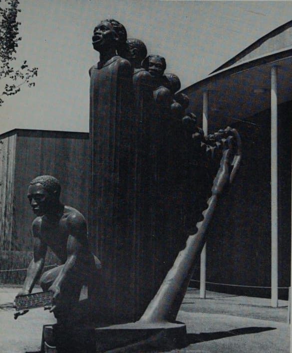 A black-and-white photograph of a large outdoor sculpture seen from the left front. A row of singing figures rises in descending height, their bodies drawn into long fluted columns like harp strings, the tallest at the left with his mouth wide open. A long tapering forearm sweeps up from the right of an oval base and ends in an open hand whose fingers curl over the smallest heads. In front, a young man kneels bare-chested with both arms extended, holding a flat ruled slab of music. Behind the sculpture stand a dark pavilion wall, a slender white column, a rope barrier on stanchions and paving, with tree branches at the top left