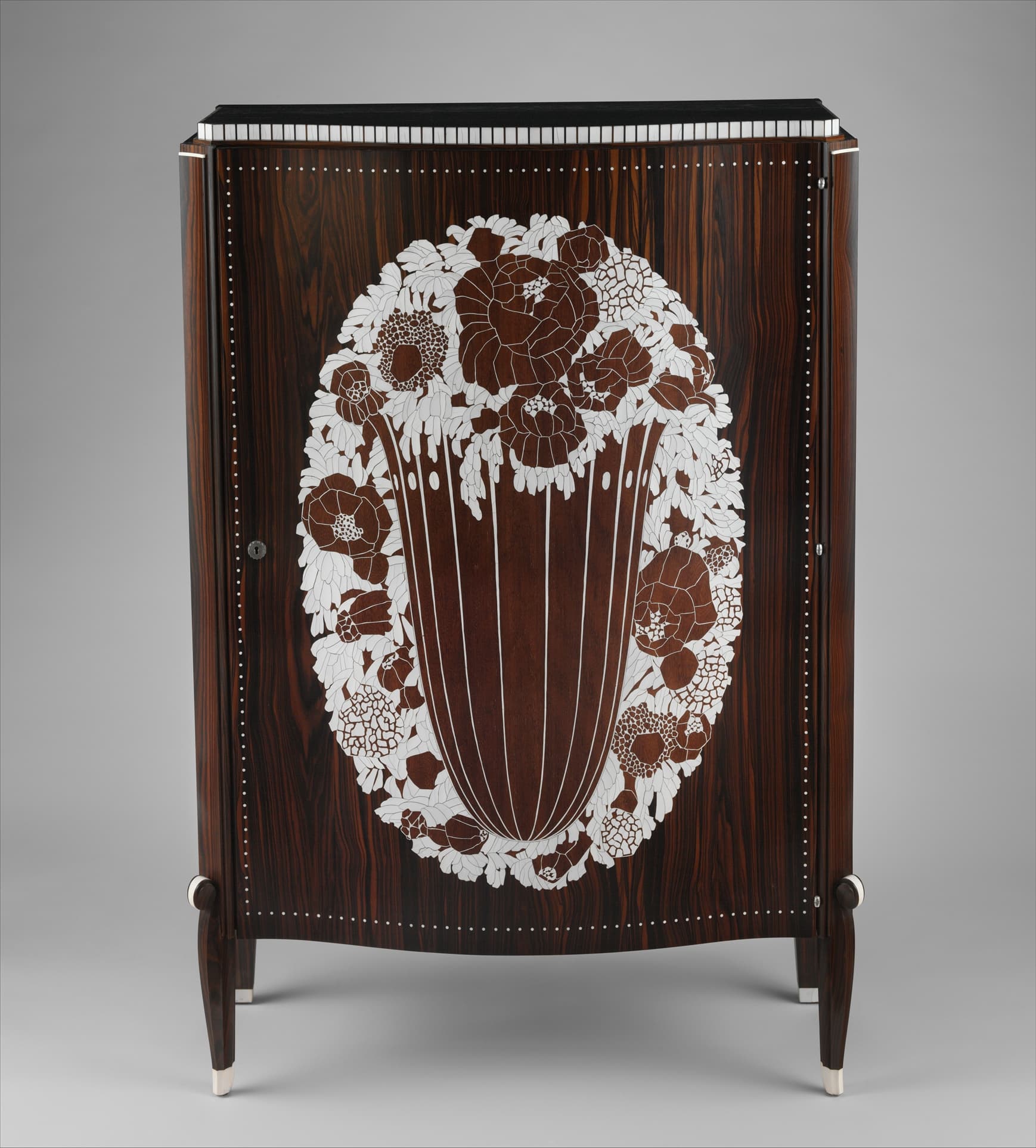 A tall dark cabinet on four slender tapering legs. The front carries a large oval of pale ivory inlay: stylised flowers massed above a tall fluted urn. A band of short ivory bars runs along the top edge, lines of small ivory dots frame the doors, and the feet are capped in ivory.