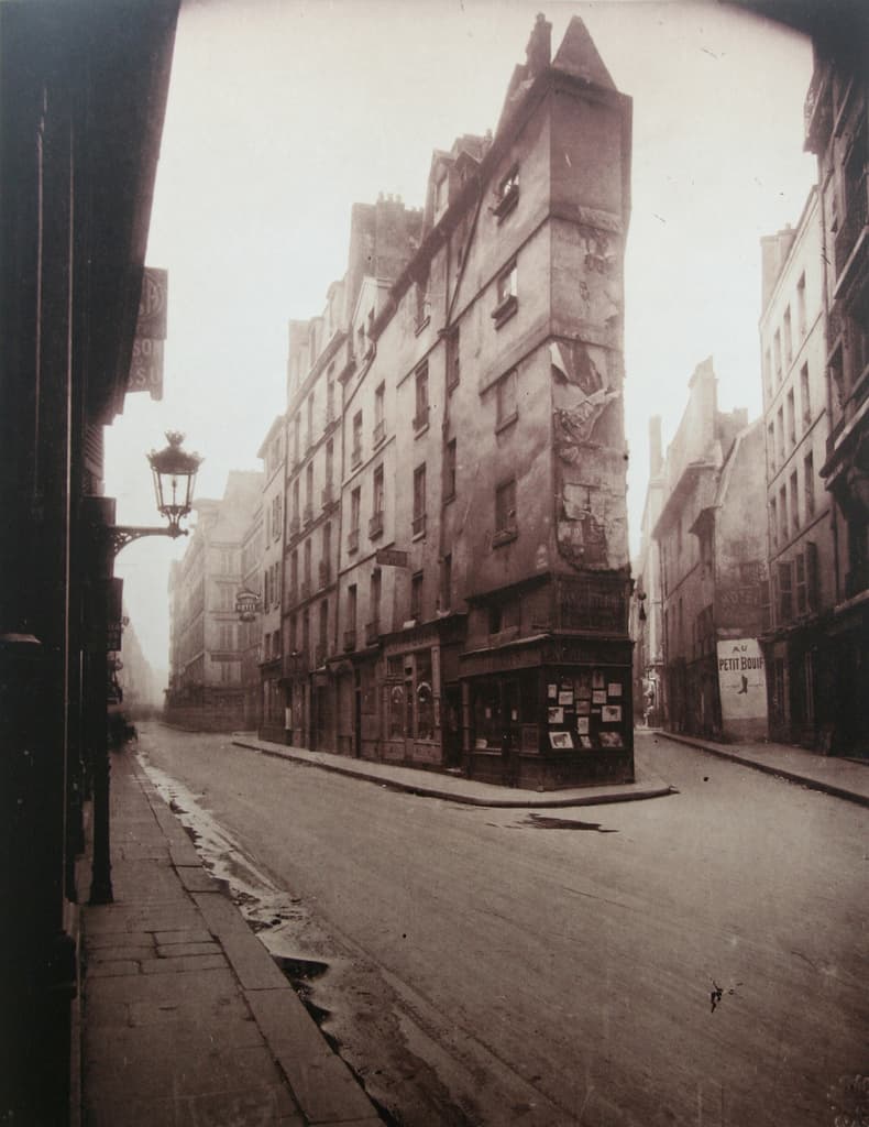 An empty narrow Paris street corner in early light, with shuttered ground-floor shopfronts, tall shuttered windows above, a wall-mounted lamp bracket, and wet paving stones receding into shadow