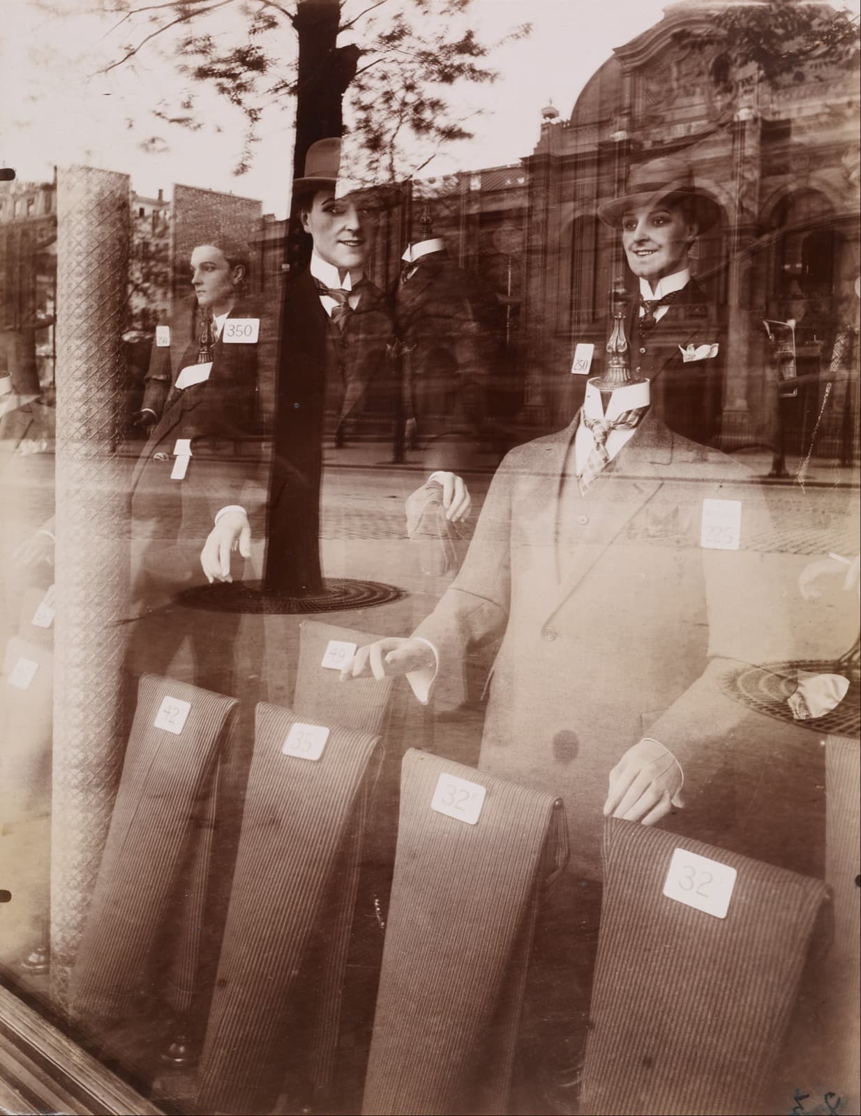 Male mannequins in suits and hats crowded behind a plate-glass shop window, with buildings and sky from the opposite side of the street reflected across the glass in front of them