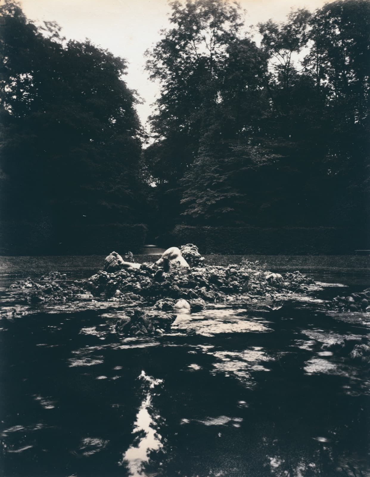 A sculpted giant's head and outstretched arm emerging from a pile of rocks at the centre of a formal garden basin, with clipped trees behind and bare branches overhead