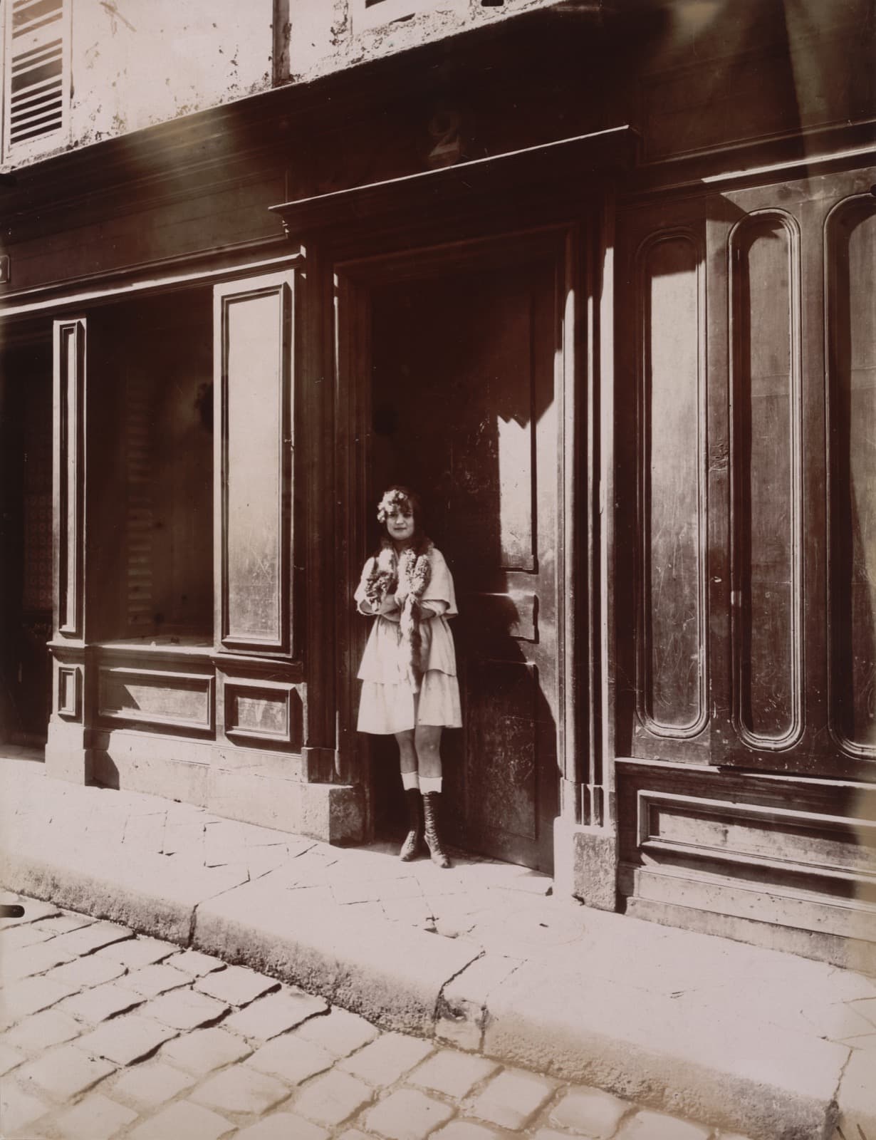 A woman in a dark dress and apron standing in the open doorway of a plain house, one hand on the frame, with a large painted street number on the wall above the door