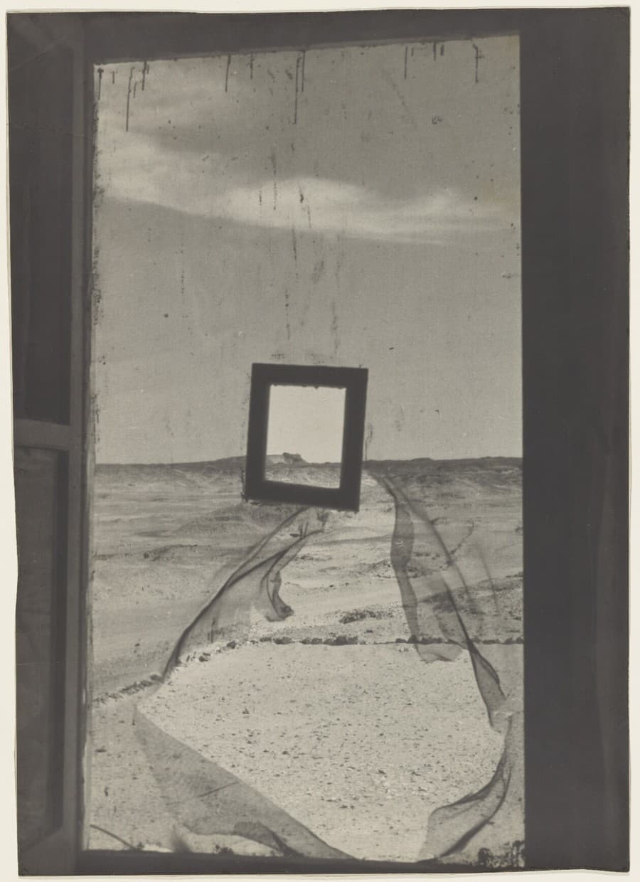 A black and white photograph of a tall opening with a dark upright at each side, filled by a fine mesh screen torn open across the middle. Through the tear a stony desert plain runs to a low ridge on the horizon under a single band of cloud. Below the tear the same desert shows through the intact mesh, greyed and flattened. An empty dark picture frame hangs in front of the screen above the tear, enclosing pale sky, the horizon and a small dark block on the ridge. Dark drip marks run down through the sky from the top edge of the print