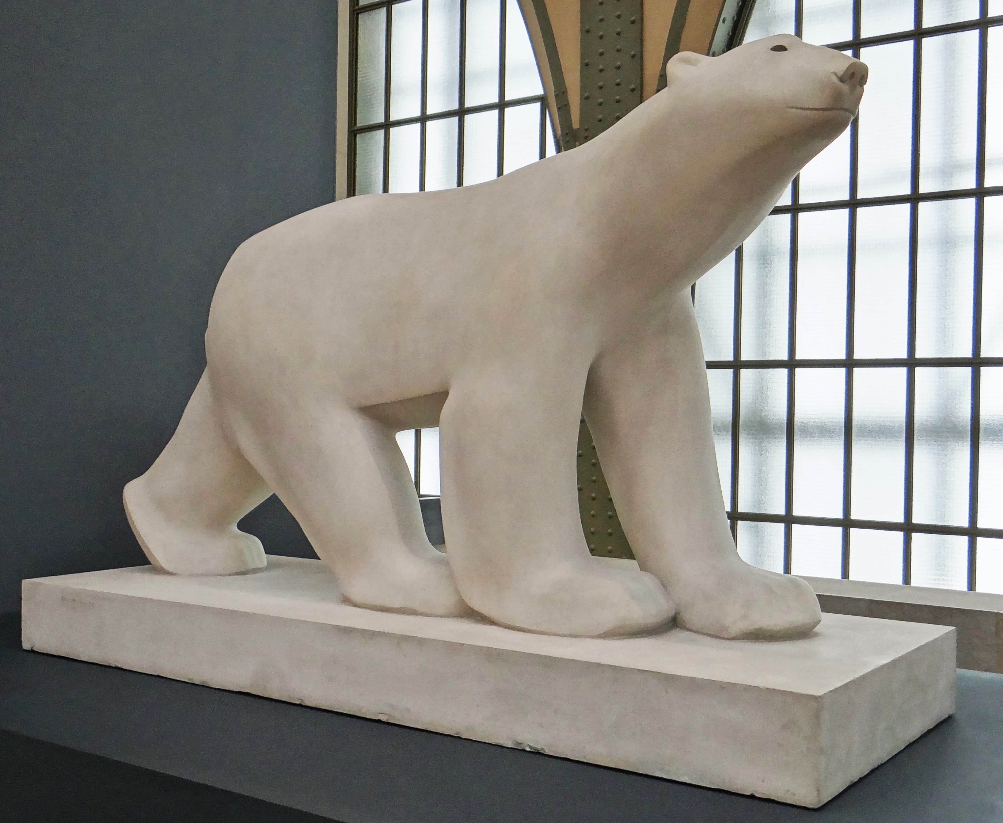 A large pale stone polar bear in profile on a plain rectangular plinth, walking with its head raised and turned slightly up. The surface carries no fur or claws: the back is one unbroken curve, the paws are rounded blocks, and the eye and mouth are shallow incised marks.