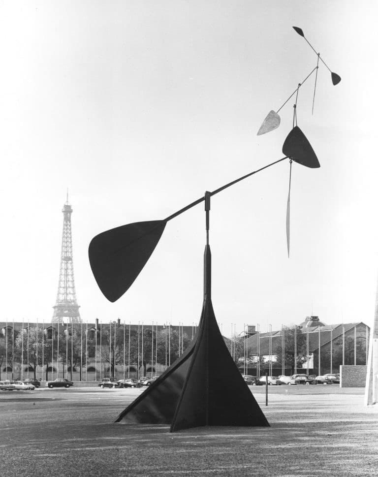 Spirale: a tall standing mobile with a dark supporting pylon and a spiralling upper arm of paddle-shaped blades, photographed against the sky in Paris