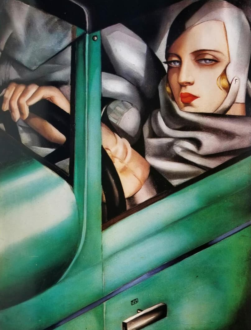 A woman at the wheel of a green car, seen through the open window. She wears a grey leather motoring helmet, a gold earring and a heavy grey scarf wound at her neck; her mouth is painted red and she looks sideways out of the frame. One gloved hand rests on the steering wheel. The car's green bodywork fills the lower half.