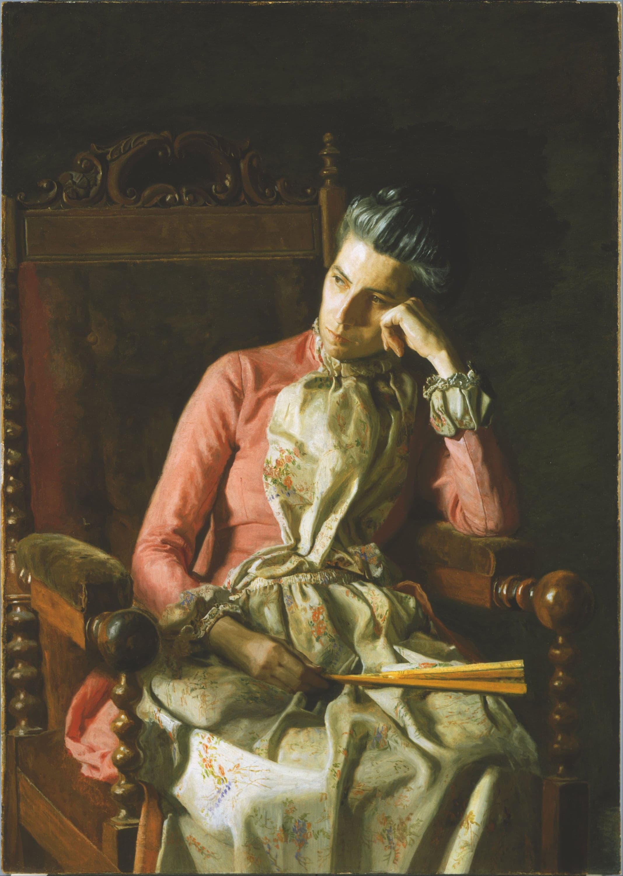 A woman with greying hair pulled back sits in a heavy dark wooden chair with a carved crest rail and turned posts, her head resting on her left hand and her eyes turned to the viewer's left. She wears a coral-pink bodice with long sleeves and a ruffled collar over a cream dress printed with small orange, green and blue flowers. Her right hand rests in her lap holding a folded yellow fan. Light falls from the left across her face and hand, and the background is dark.