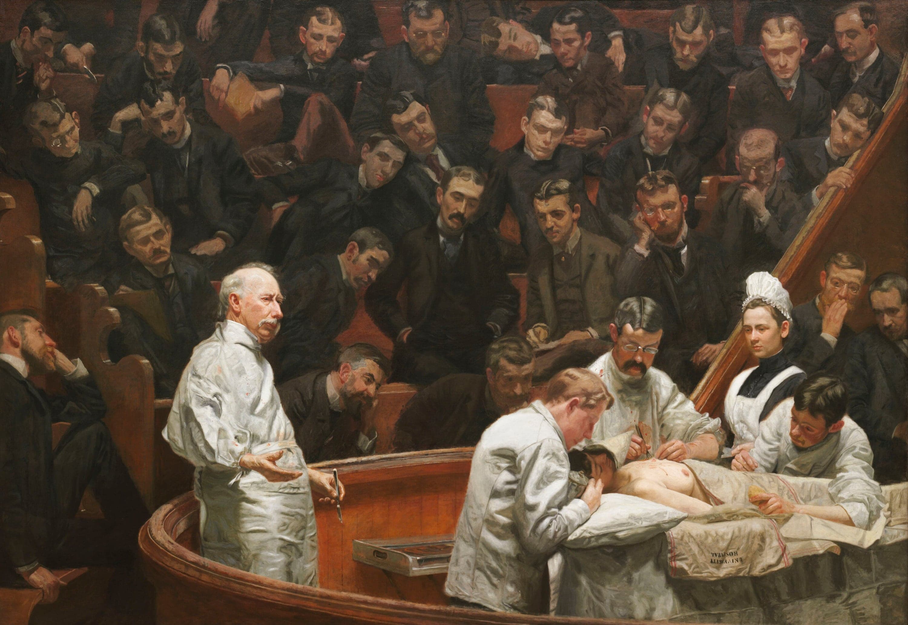 A wide painting of a surgical amphitheatre with red-brown wooden benches. At the left an elderly surgeon with white hair and a white moustache stands in a white gown holding a scalpel loosely in his right hand, his left hand open as he speaks. At the right three doctors in white work over a woman lying on a table with her chest bare, one bending over her head, one in spectacles at the incision, one holding her arm, while a nurse in a white cap and black dress stands behind them. Rows of young men in dark suits fill the benches above, and a metal instrument case sits on the rail in front of the surgeon.