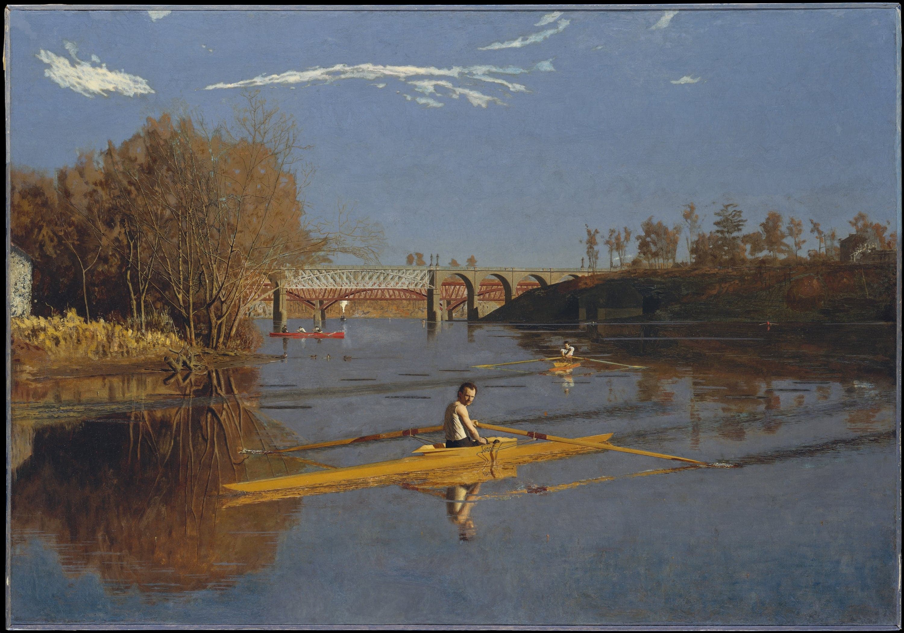 A wide river scene under a blue sky with thin white clouds. In the foreground a dark-haired man with a moustache, in a white sleeveless shirt, sits in a pale yellow racing scull with his oars trailing and looks back over his shoulder. A second rower in a white shirt pulls away in an orange-red scull in the middle distance. Beyond them a red boat with three rowers and a small steam launch sit under an iron truss bridge on stone piers, with a stone bridge of round arches behind it. Bare autumn trees and the corner of a stone house stand on the left bank, and the still water reflects everything.
