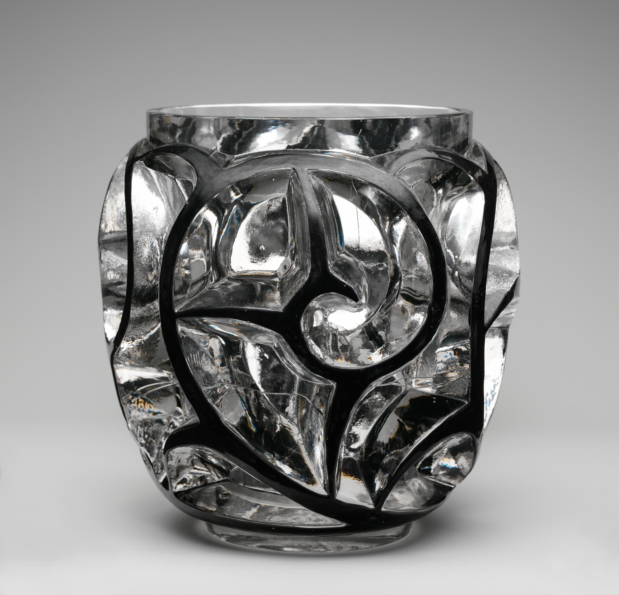A squat clear glass vase, wider at the shoulder than at the foot. Thick curling forms are cut deep into the wall and sweep around it, one spiralling inward at the centre. Black enamel fills the recessed channels between them. The neck and the foot are left smooth.