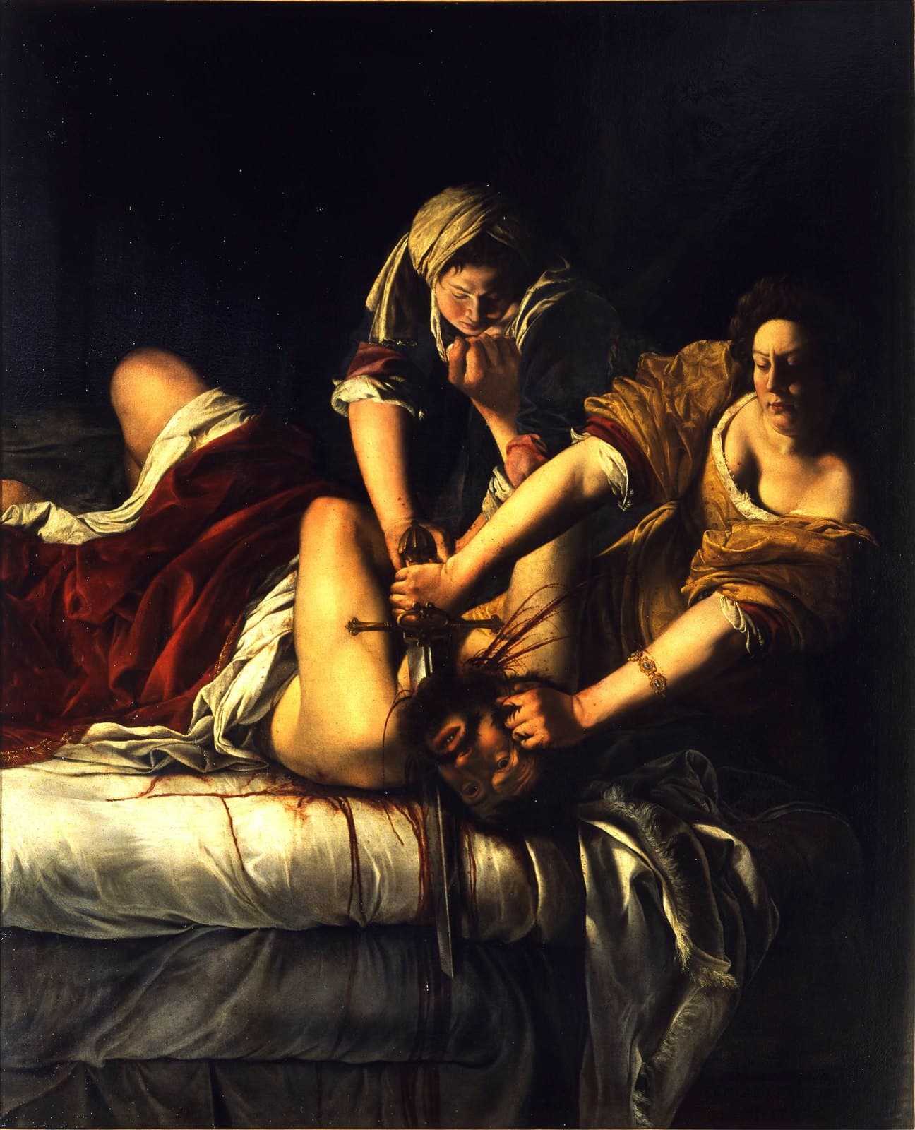 Two women lean over a bearded man lying on a white-sheeted bed. The younger woman in a golden-yellow dress grips his hair with one hand and drives a long sword into his neck with the other; a second woman in blue-grey pins his chest with both hands while he pushes up at her. Blood arcs from the neck and runs down the sheets, and the background behind them is almost black.