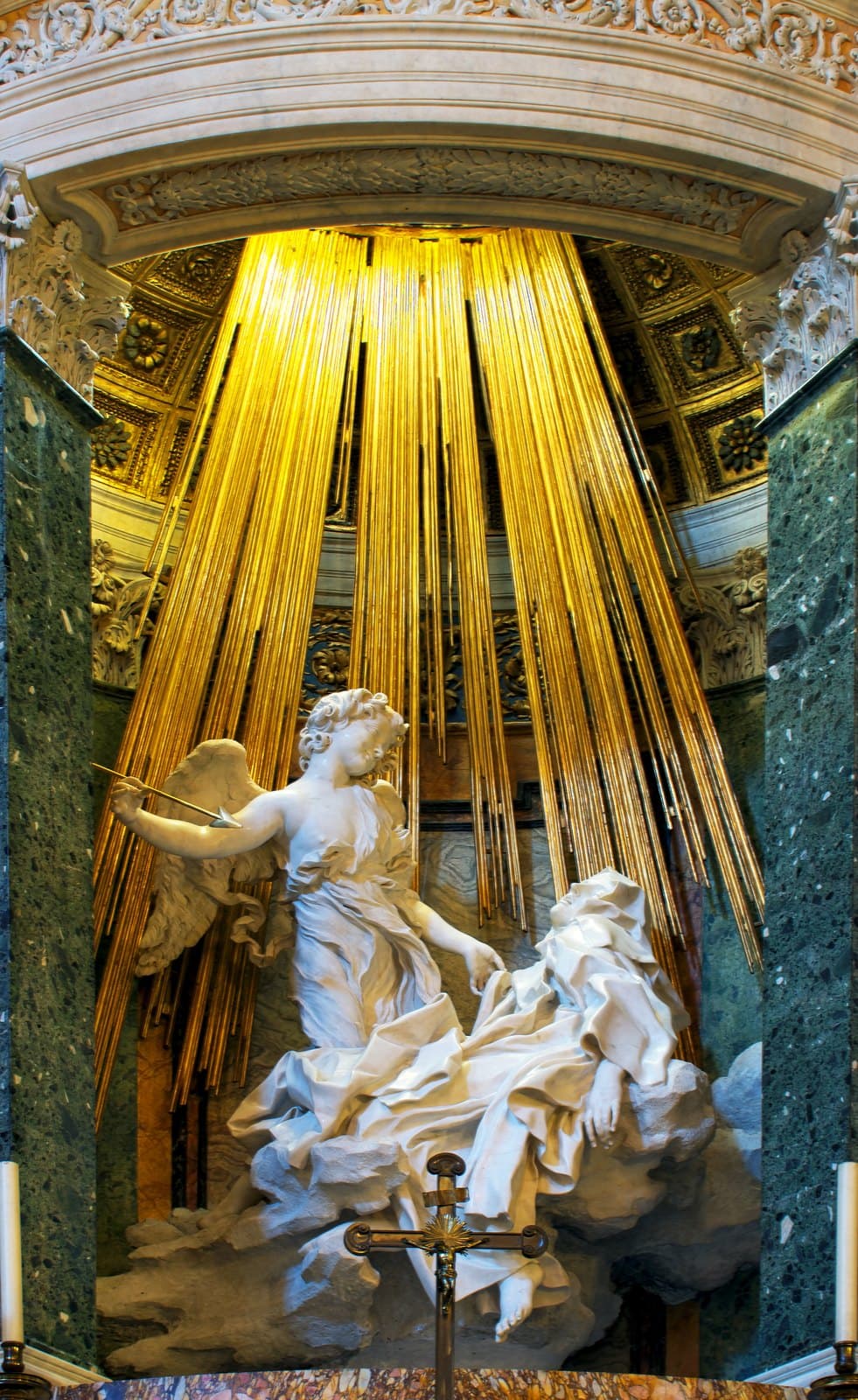 White marble group in a chapel niche framed by dark green marble columns: a winged angel in a loose robe stands at the left holding a golden arrow with a metal point, his other hand lifting the edge of the habit of a nun who has fallen back on a marble cloud with her eyes closed, her mouth open and one bare foot hanging down. Long gilded rods hang behind them from an opening high above.