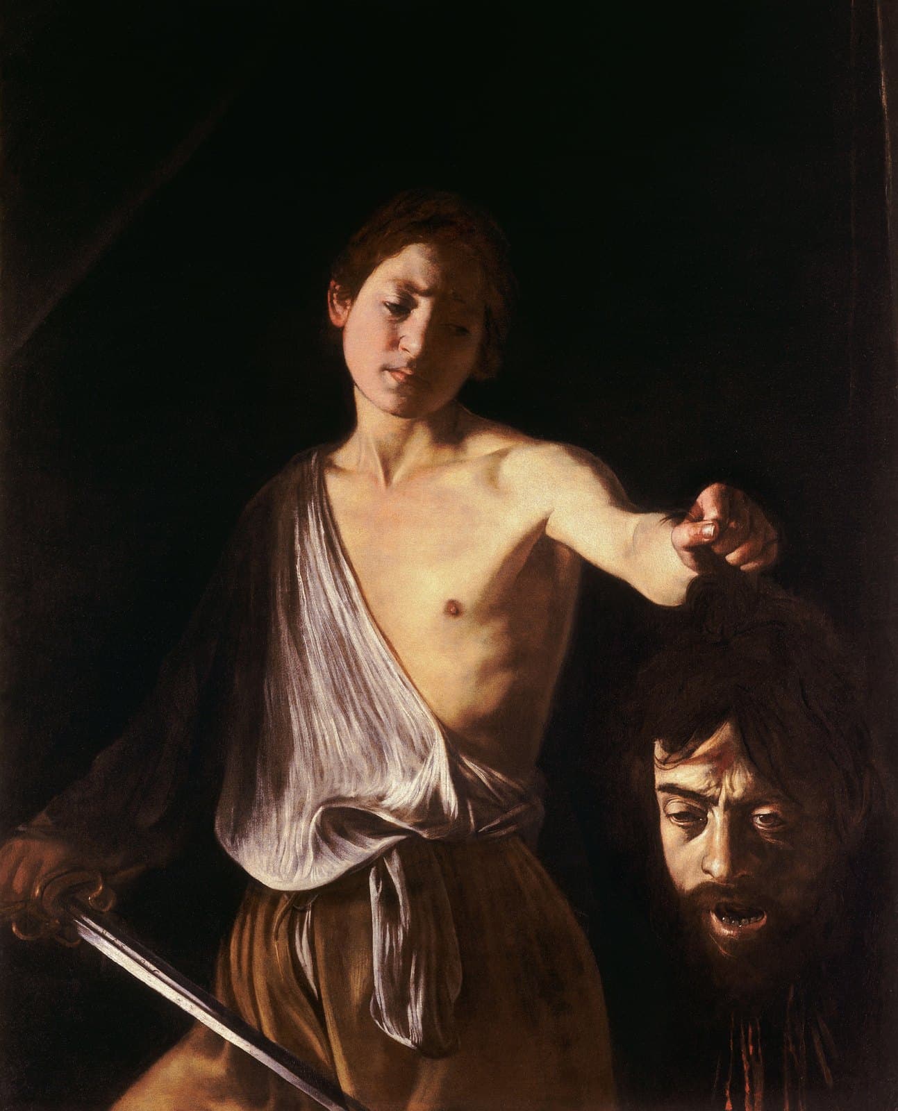 A bare-shouldered young man in a loose white shirt stands against a black background, a sword in his lowered right hand. With his left arm outstretched he holds a severed bearded head up by the hair. The head's mouth hangs open and its eyes are half shut. The young man looks down at it without triumph