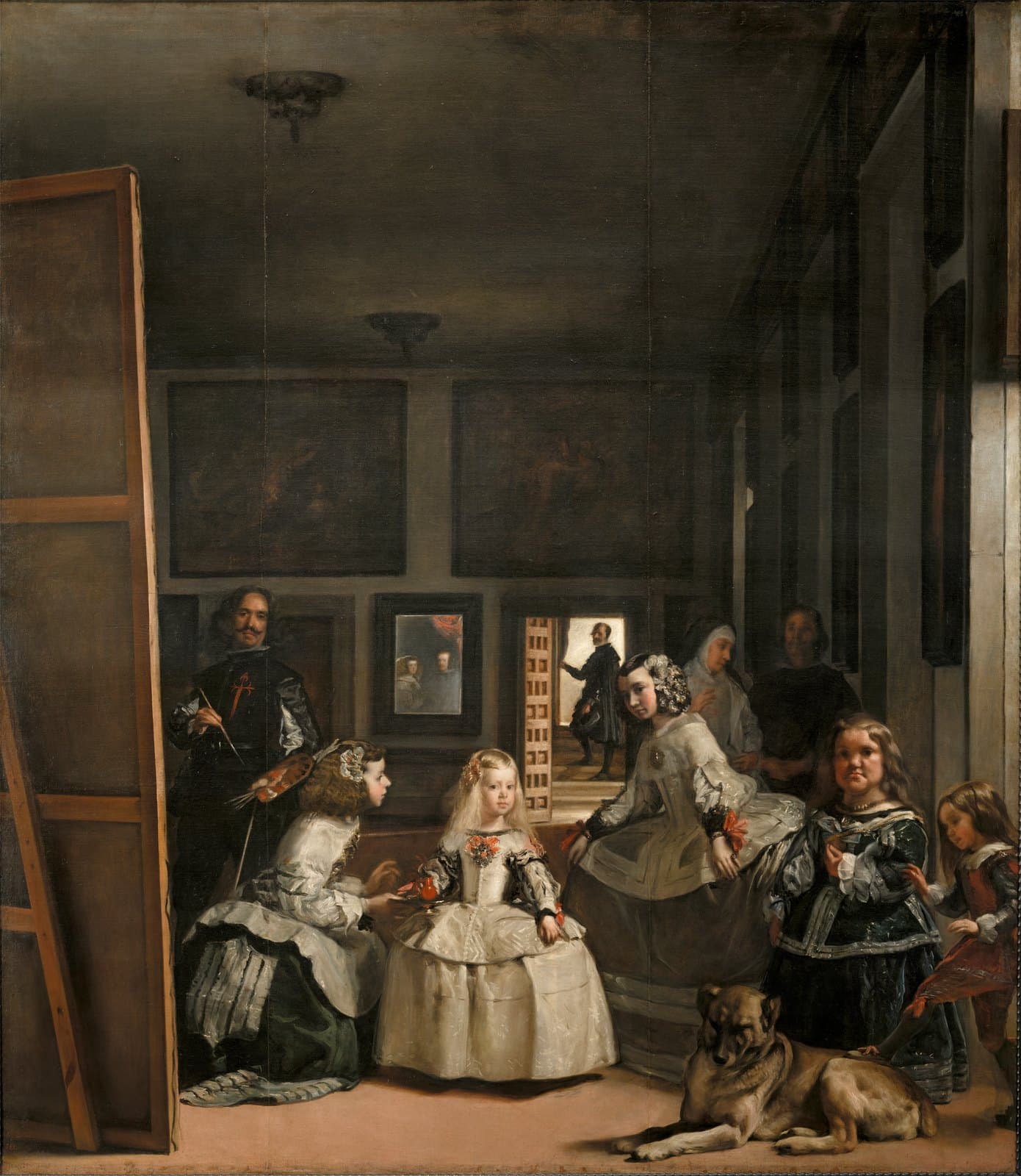A tall, dim palace room. A small blonde girl in a wide pale gown stands near the center; a young woman kneels at her left offering a red cup on a tray, and a second young woman leans in from her right. Further right, a stocky woman in dark green and a boy who rests his foot on a large sleeping mastiff. To the left the painter stands at the back of a huge canvas, brush and palette in hand, a red cross on his black doublet. Behind them a small mirror on the back wall holds two lit faces, and a man stands in a bright doorway on a flight of steps.