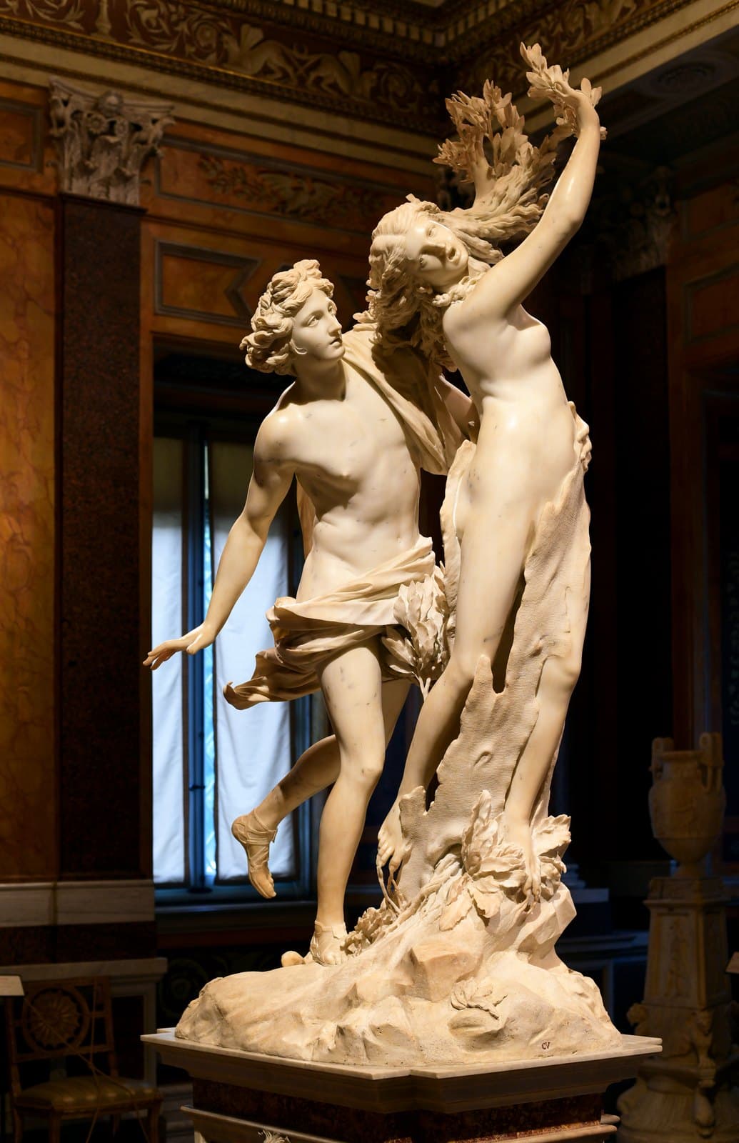 White marble group on a pedestal in a gallery: a running young man in a flying cloak reaches around the waist of a woman whose raised hands are sprouting laurel branches, whose hair has turned to leaves and whose legs are being wrapped in bark and roots