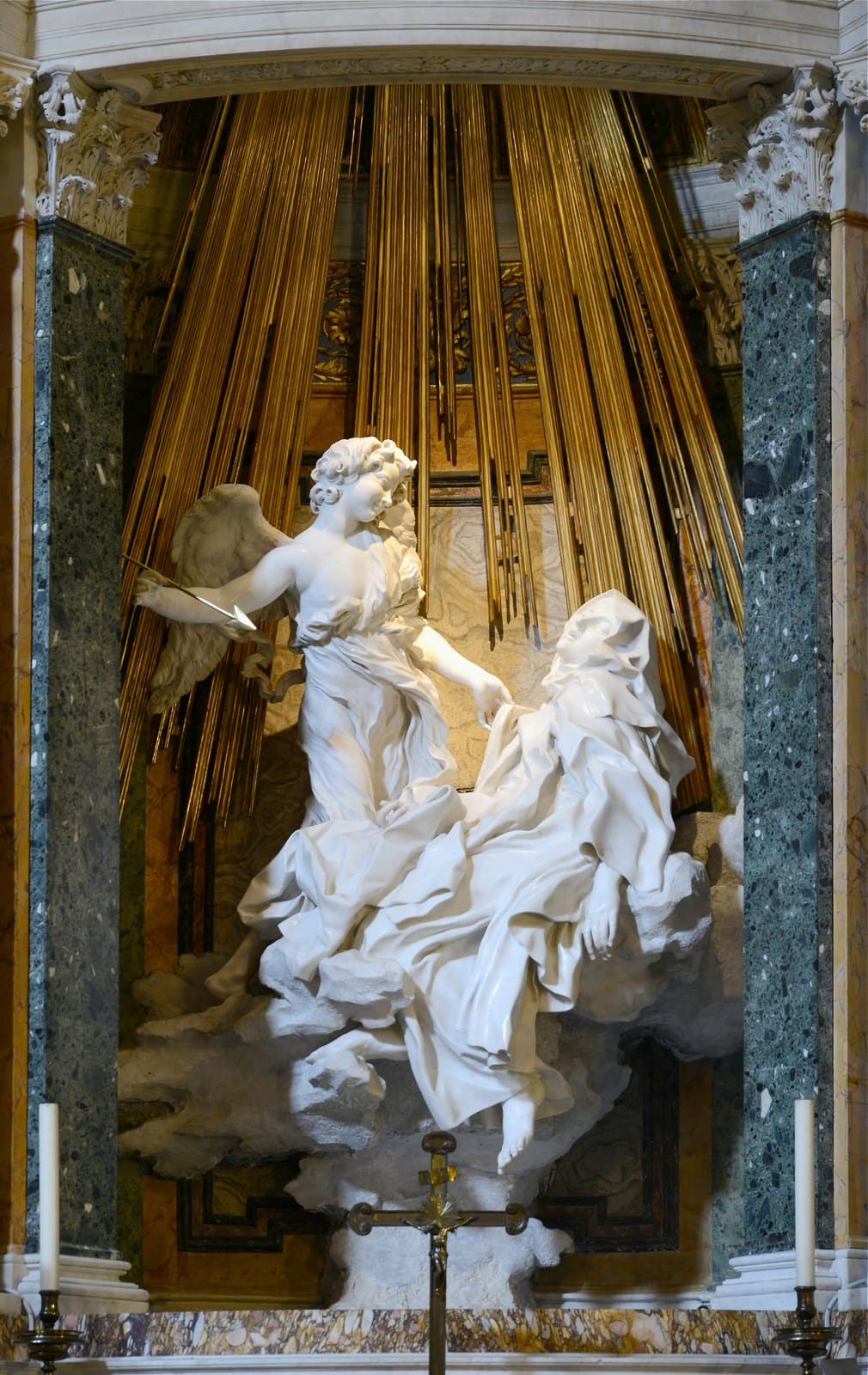 White marble group set in a niche between green marble columns: a smiling winged youth holds a golden arrow and lifts the robe of a nun who has fallen back on a marble cloud with her eyes shut, her mouth open and one bare foot hanging down, with gilded rods behind them standing for falling light
