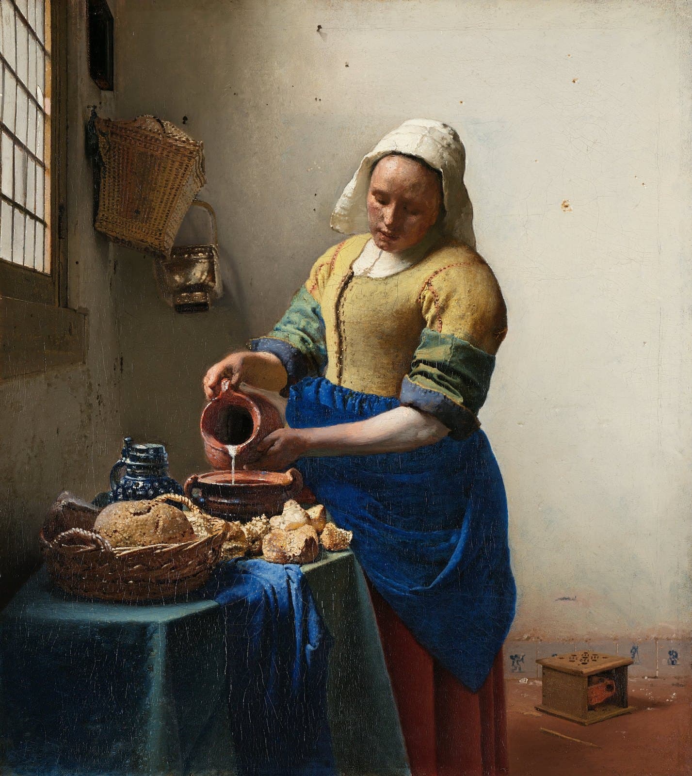 A young woman in a white cap, yellow bodice and deep blue apron stands at a table beside a leaded window, tipping a brown earthenware jug so that a thin stream of milk falls into a wide pot. A basket of bread, loose crusts, a blue-grey stoneware jar and a woven basket sit on the blue-draped table, and a small wooden foot warmer stands on the tiled floor behind her.