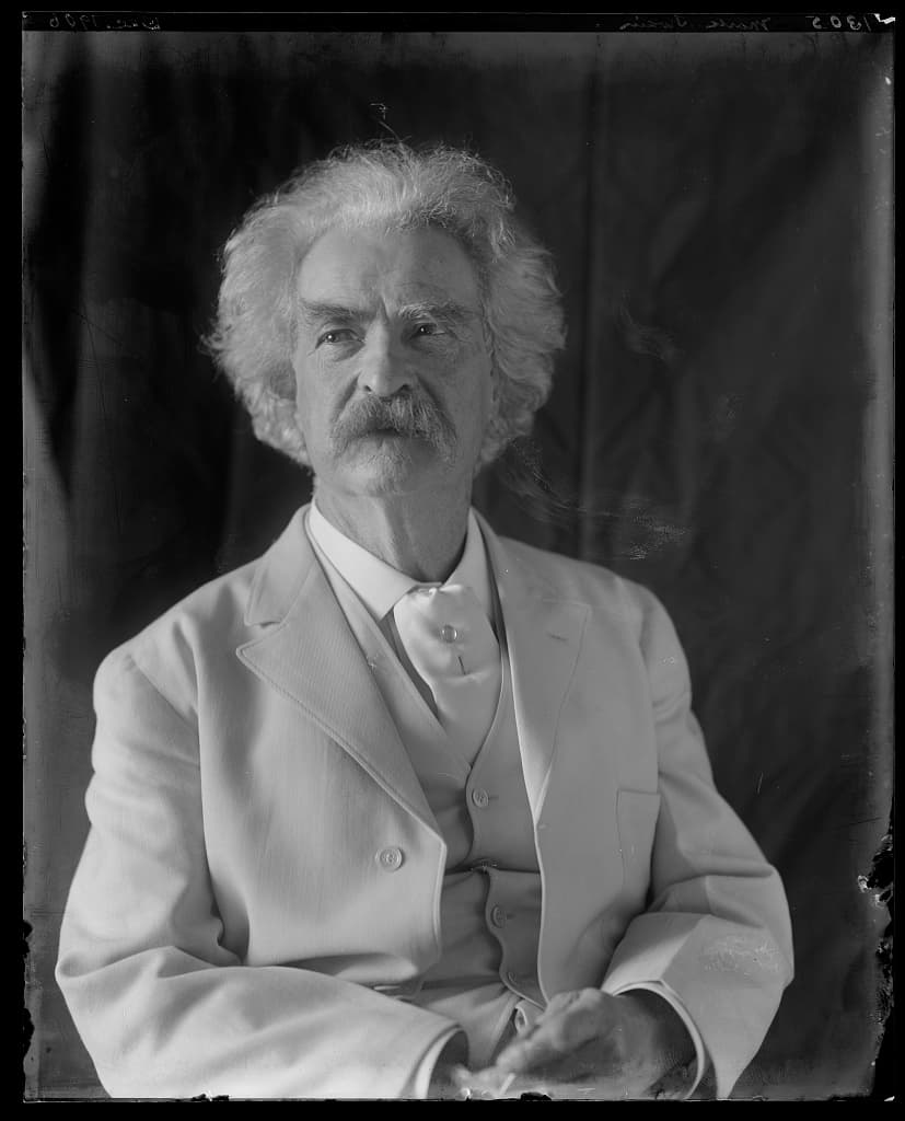 Mark Twain seated in a light suit for photographer Frances Benjamin Johnston in 1906, with his white hair and moustache brightly lit against a dark ground