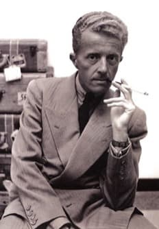 Paul Bowles in a light suit and dark tie, seated in front of stacked steamer trunks, holding a cigarette and looking at the camera