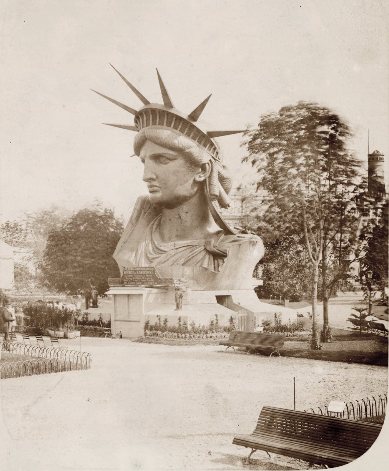 The copper head and shoulders of the Statue of Liberty standing on a low plinth in a Paris park, spiked crown against the sky, with gravel paths, park benches, young trees and two small figures beside the base for scale
