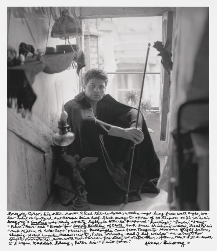 A black and white photograph of a young man sitting on the floor in the corner of an attic, knees drawn up, wrapped in a dark coat with a high collar, looking straight at the camera. A long thin rod crosses his body towards an open casement window at the right, where the daylight has burned out to blank white. A shelf above his head carries a toy bull, a round tin and a hanging doll in a full skirt, and a glass oil lamp stands against the wall at his shoulder. Eight lines of handwriting in ink run across the white border below the picture, signed Allen Ginsberg