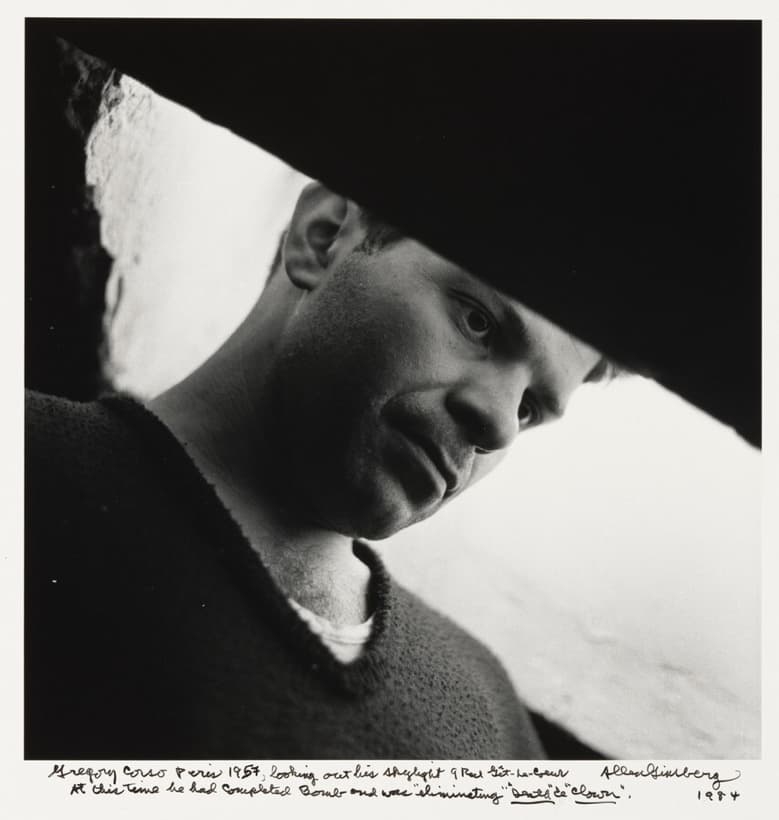 A black and white close-up of a man's face lying diagonally across the frame, lit hard from one side, with several days of stubble and one open eye near the centre carrying a catchlight. A solid black wedge with no detail fills the upper right and a blown white wedge runs along the lower right edge. He wears a dark crewneck sweater. Two lines of handwriting in ink run along the white border below, signed Allen Ginsberg and dated 1984 at the right