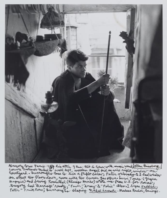 A black and white photograph of a young man sitting on the floor of an attic in a dark coat, head tipped back and turned away from the camera, his raised left hand gripping a long thin rod that stands almost vertical. A glass oil lamp stands at his left shoulder and a shelf above carries a toy bull, a round tin and a hanging doll in a full skirt. An open casement window at the upper right shows a courtyard burned almost to white. Nine lines of handwriting in ink fill the white border below, with a signature boxed inside a drawn rectangle at the end of the first line
