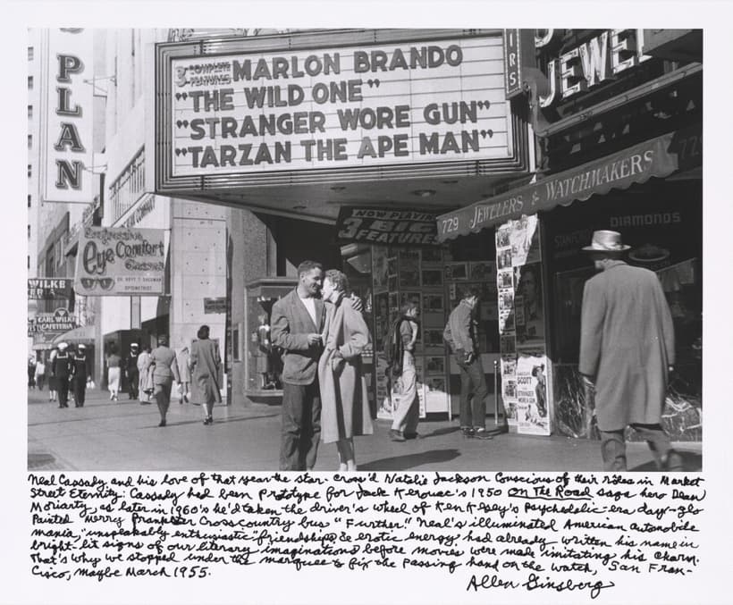 A black and white street photograph. A man in a light tweed jacket, white T-shirt and dark jeans holds a laughing woman in a pale full-length swing coat under a cinema marquee reading 3 COMPLETE FEATURES, MARLON BRANDO, THE WILD ONE, STRANGER WORE GUN, TARZAN THE APE MAN. A wall of glazed film posters covers the cinema front behind them and a scalloped awning at the right reads 729 JEWELERS AND WATCHMAKERS. A man in a pale overcoat and light hat walks away from the camera at the right edge, out of focus, and sailors in white caps walk up the block at the far left. Eight lines of handwriting in ink fill the white border below, signed Allen Ginsberg on a ninth