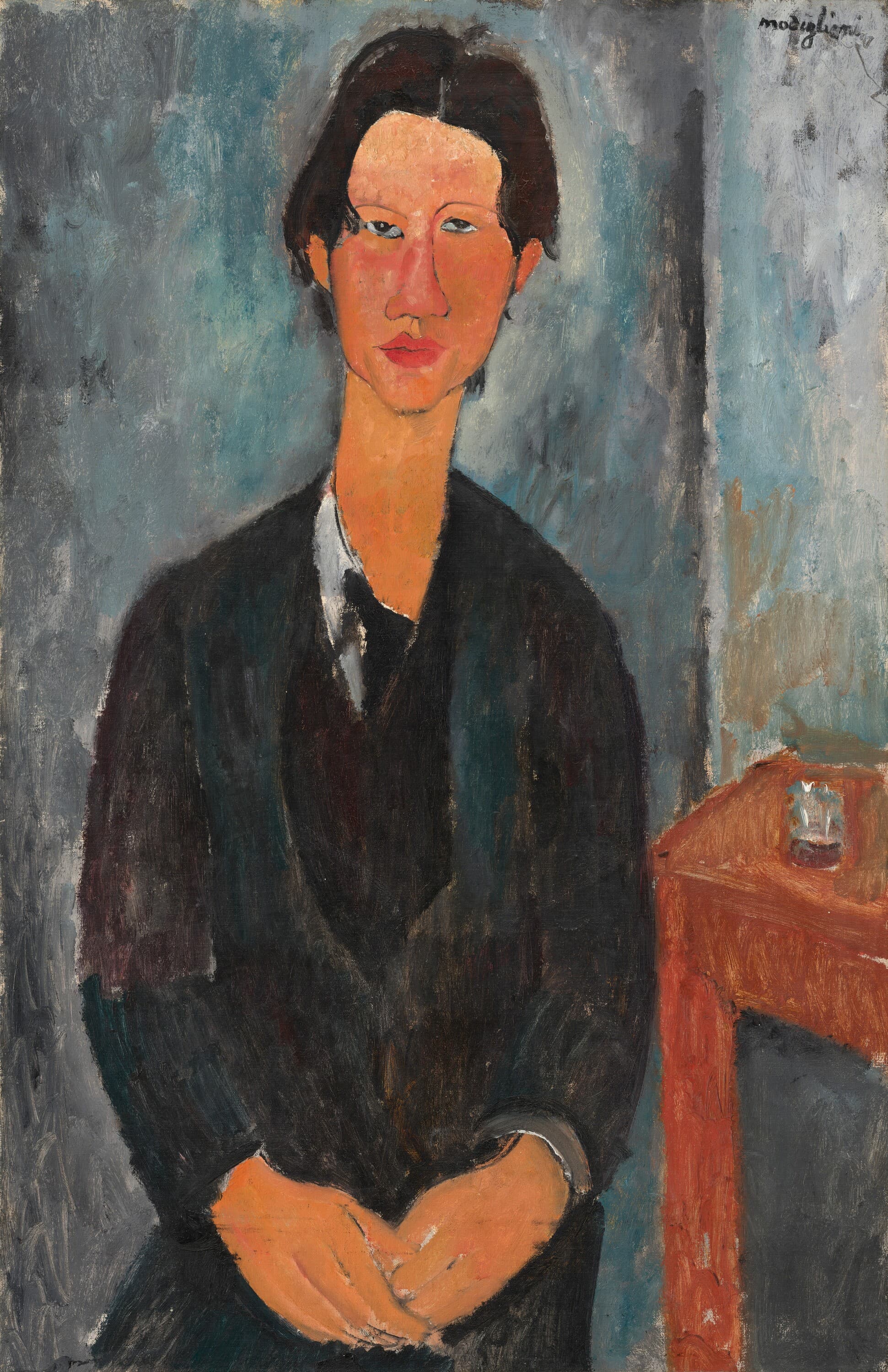 A young man with black hair parted in the middle sits facing us, his long face slightly lopsided, with gray-blue eyes and full red lips. He wears a black coat over a dark green waistcoat and a white collar, and his hands rest together in his lap. A small red table with a glass stands at the right against a gray-blue wall. Signed modigliani at upper right