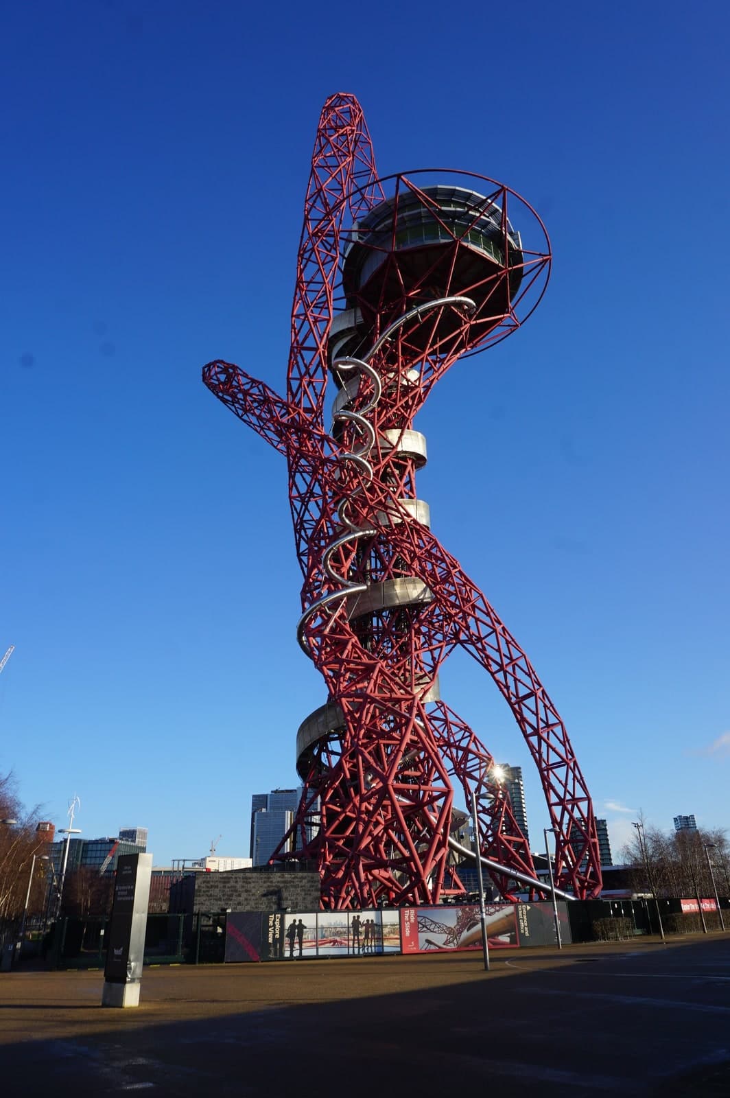 A tall red steel lattice tower photographed from ground level against a cloudless deep blue sky. Loops of red tubular steel wind around and across each other, one arm swinging out to the left and another curving down to the right, with a tight vertical loop rising above the top. A grey glazed viewing pod sits near the summit, ringed by a red hoop. A bright silver tube spirals down the outside of the structure through the red steelwork. At the base, hoardings carrying photographs and the words Explore The View and Ride The Slide run across an empty paved forecourt, with lamp posts, bare winter trees, a construction crane and tower blocks behind