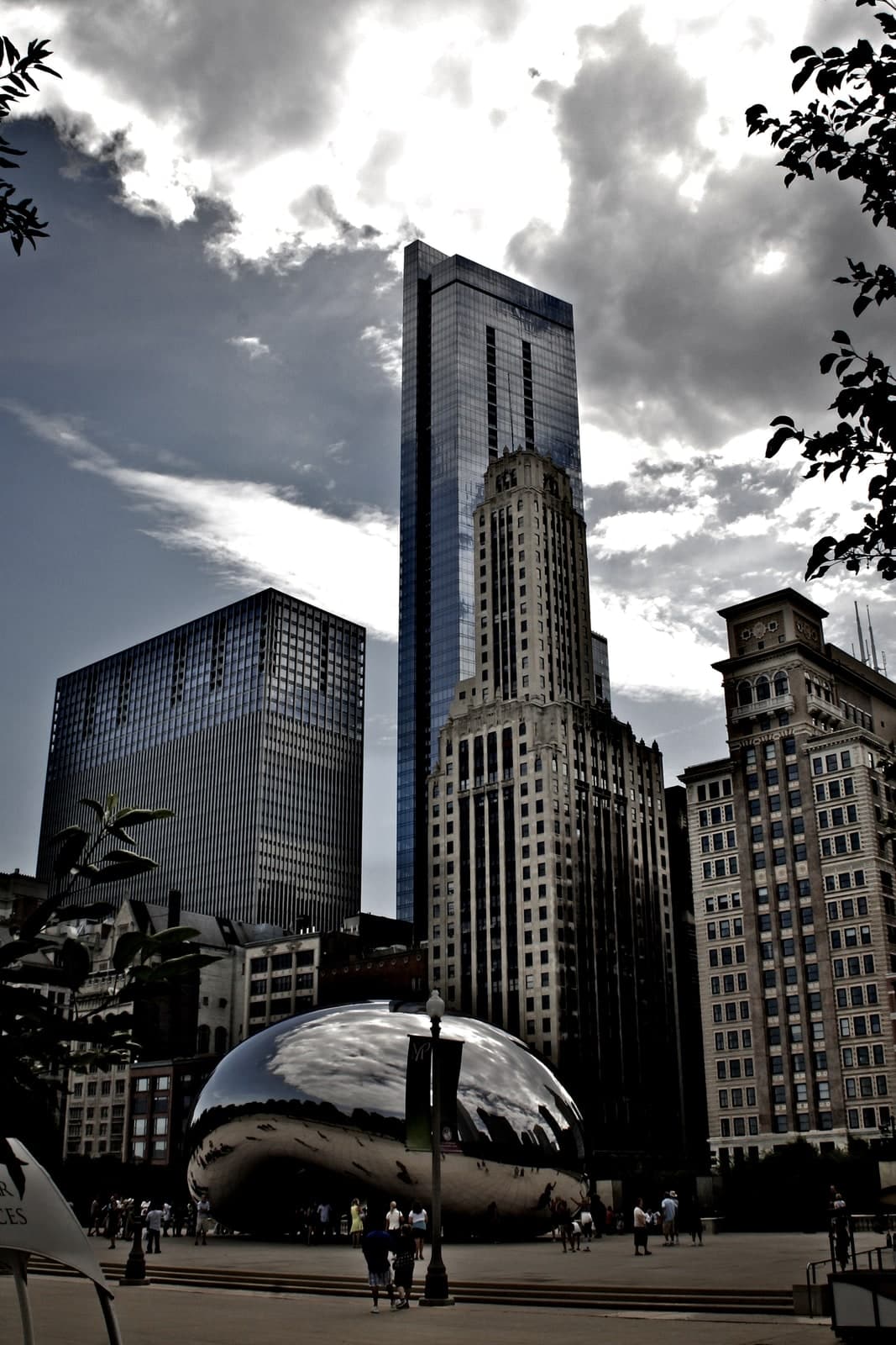A mirrored steel ellipsoid standing on a paved plaza, seen end-on from across the square under a heavily clouded sky. Its polished surface carries a compressed, curved reflection of the buildings and clouds around it, with a dark band along the lower edge. Beneath the sculpture an arch opens into a darker chamber, and small figures stand under it and along its flanks. Behind and above rise a blue glass tower, a pale stone skyscraper with a stepped crown, a dark modernist slab on the left and a brick tower on the right. A street lamp stands in front of the sculpture and leaves overhang the top corners of the frame