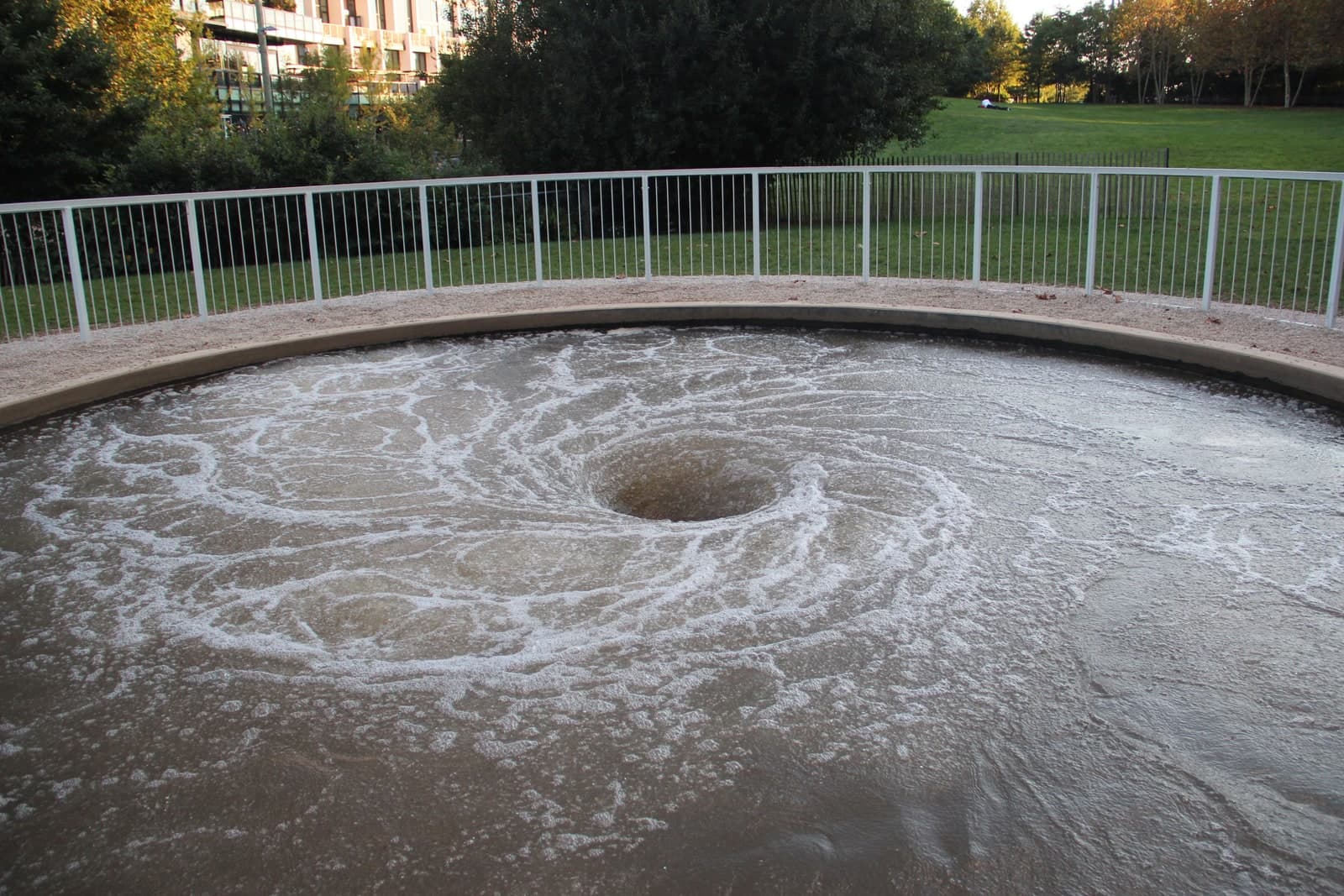 A circular pool of dark, turbulent water set into the ground and photographed from the rim. The water spins into a funnel, with a black opening at the centre and broad arms of white foam curling out from it across the whole surface. A low pale stone kerb runs round the pool, with pinkish gravel beyond it and a white metal railing set on a black fence enclosing the far side. Behind the railing are mown grass, dense green trees and, at the top left, a modern apartment building with balconies