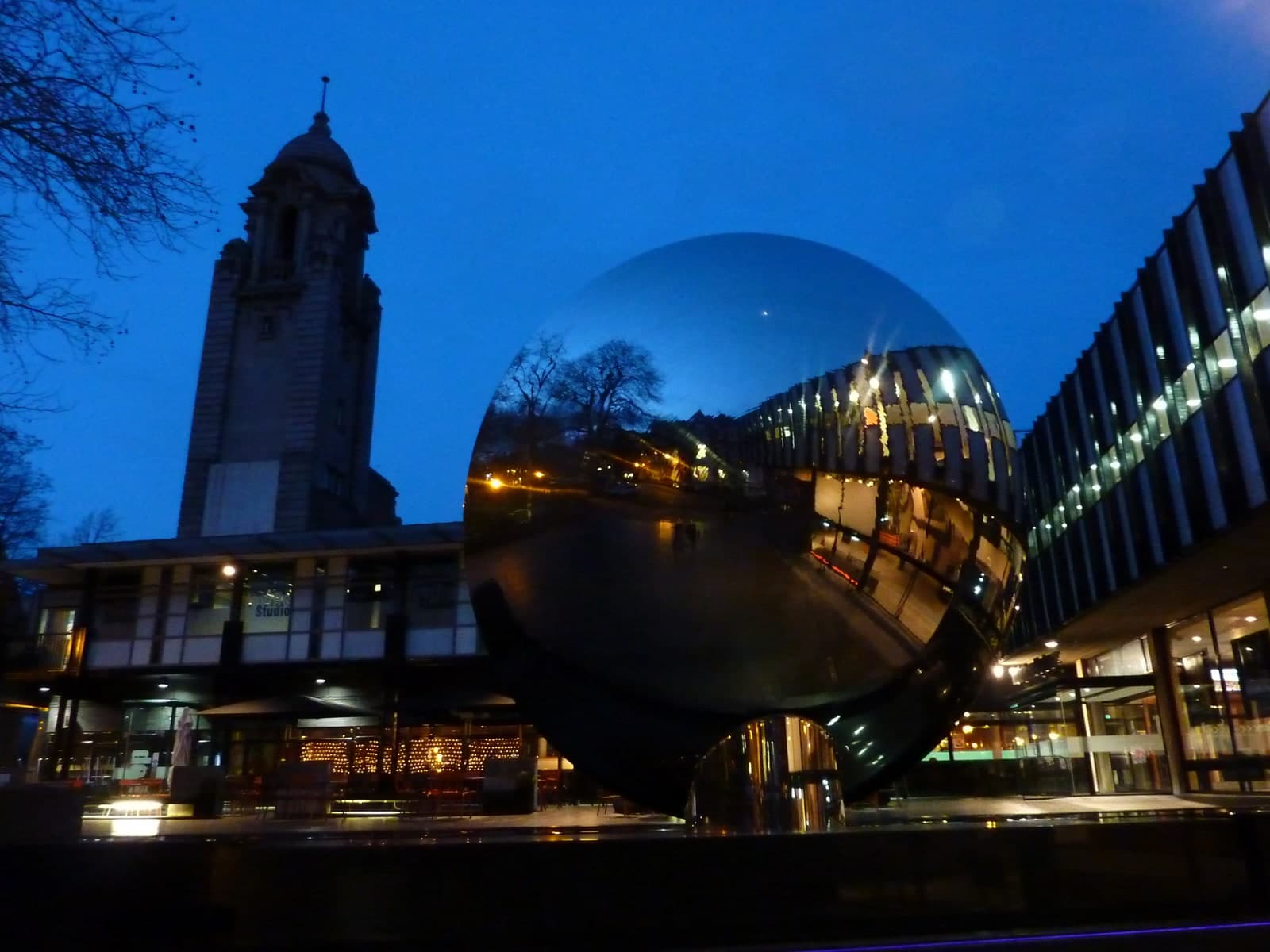 A large circular mirrored dish standing on end outside a theatre at dusk under a deep blue sky. Its concave face holds an inverted reflection: bare trees and a lit street run across the upper part of the disc, orange street lamps and a wet road below them, and the glazed front of the theatre curves round the right-hand side. The lower part of the disc is dark. Behind and to the left stands a tall stone bell tower with a domed lantern, with a low glazed theatre frontage lit from inside beneath it, and a glass and steel building to the right
