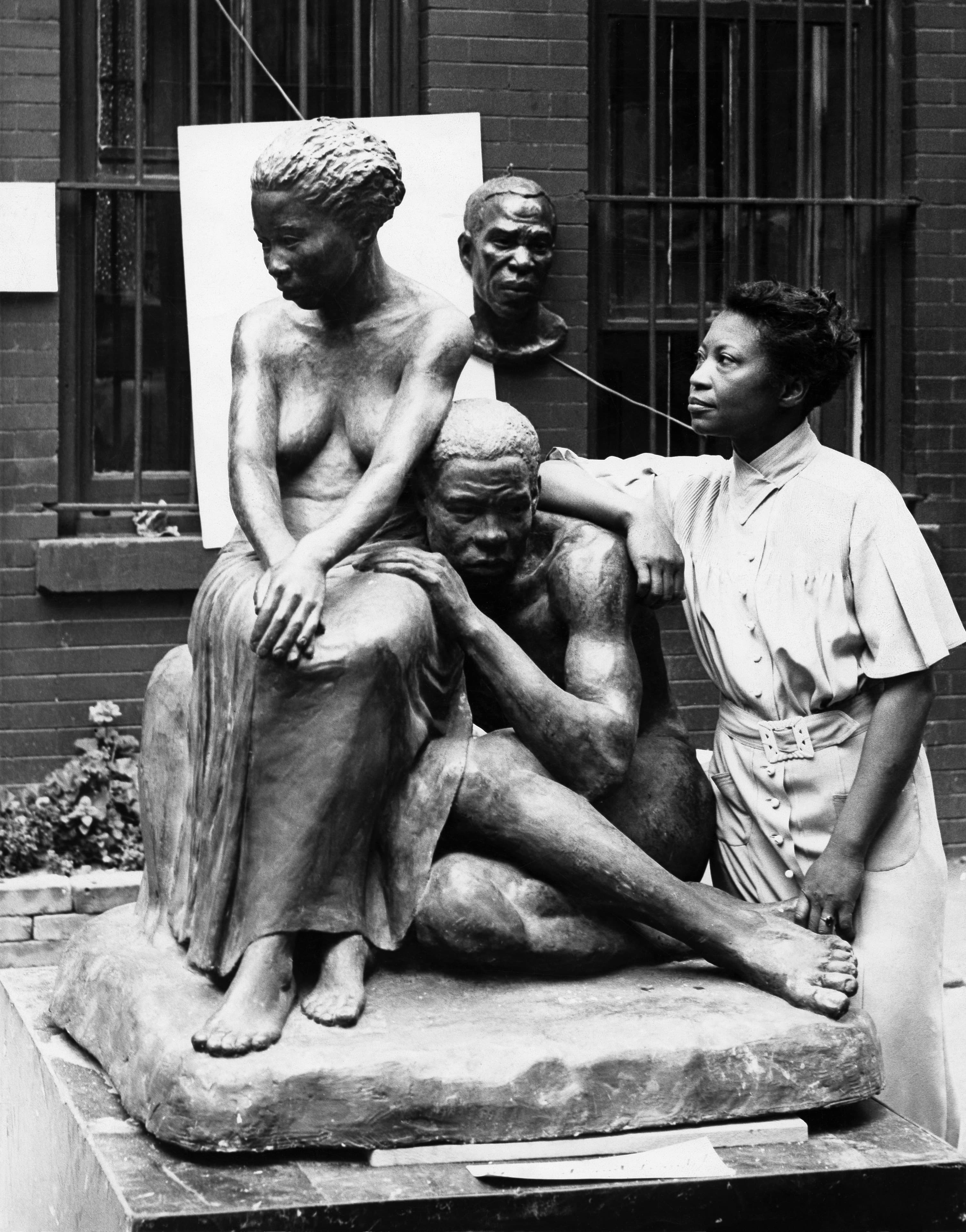 A black-and-white photograph of a large sculpture standing outdoors on a low platform against a brick wall with barred windows. Two figures sit on a rough slab shaped like an outcrop of rock: a woman upright at the left, bare to the waist with a long skirt and bare feet, her head bowed and her hands folded over her knee, and a man below her with his legs stretched out to the right, one arm hooked over her knee, his head dropped forward. Augusta Savage stands at the right in a light short-sleeved dress with a buckled belt, her left forearm resting on the man's shoulder, looking away from the camera. A separate portrait bust of an older man stands on a socle behind the group, and geraniums grow at the lower left