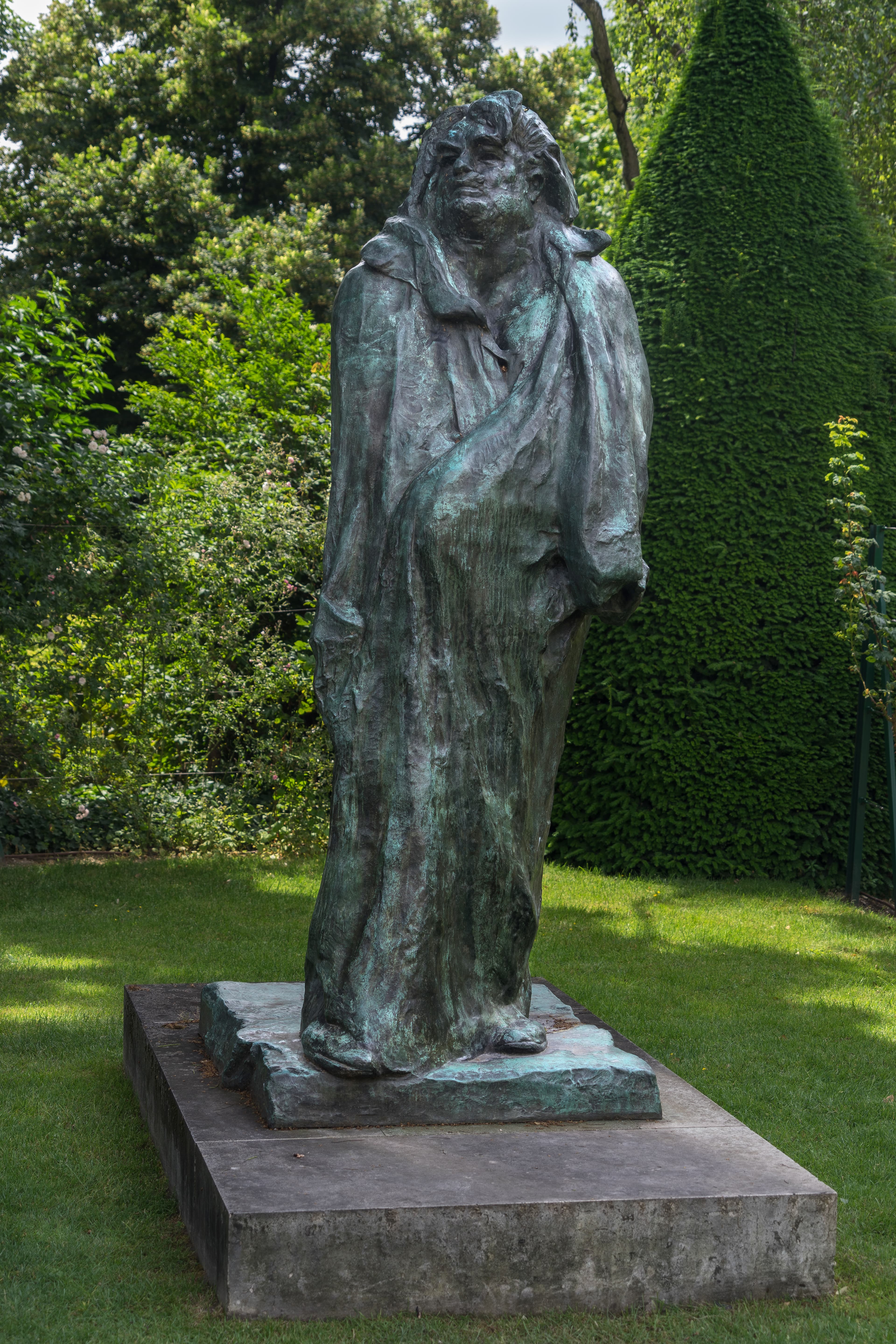 A tall bronze of a standing figure wrapped from neck to ground in a long loose robe, with no arms or hands visible. The head is thrown back, with deep-set eyes under a heavy brow and a thick mane of hair swept back into two masses. The patina is pale blue-green over grey-brown, streaked by rain. The bronze stands on a rough irregular bronze slab set on a low grey stone plinth on a lawn, with a clipped conifer and flowering shrubs behind