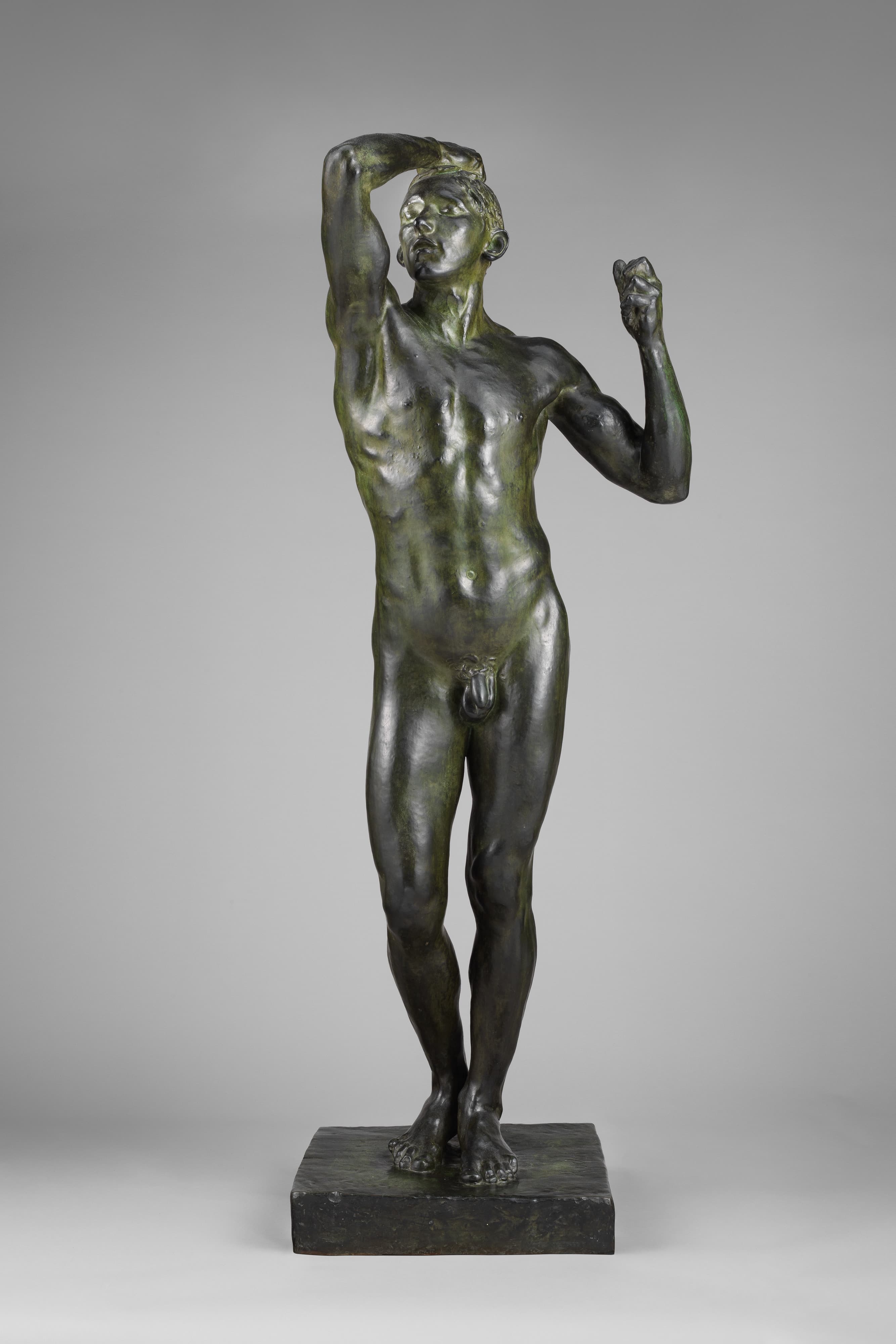 A life-size bronze of a naked young man standing with his head tipped back. His right arm is bent above him with the hand resting on his head. His left arm is raised and folded at the elbow, the fingers curled around empty air. The patina is dark brown with olive-green mottling. The figure stands on a thick rectangular bronze base against a plain grey studio backdrop