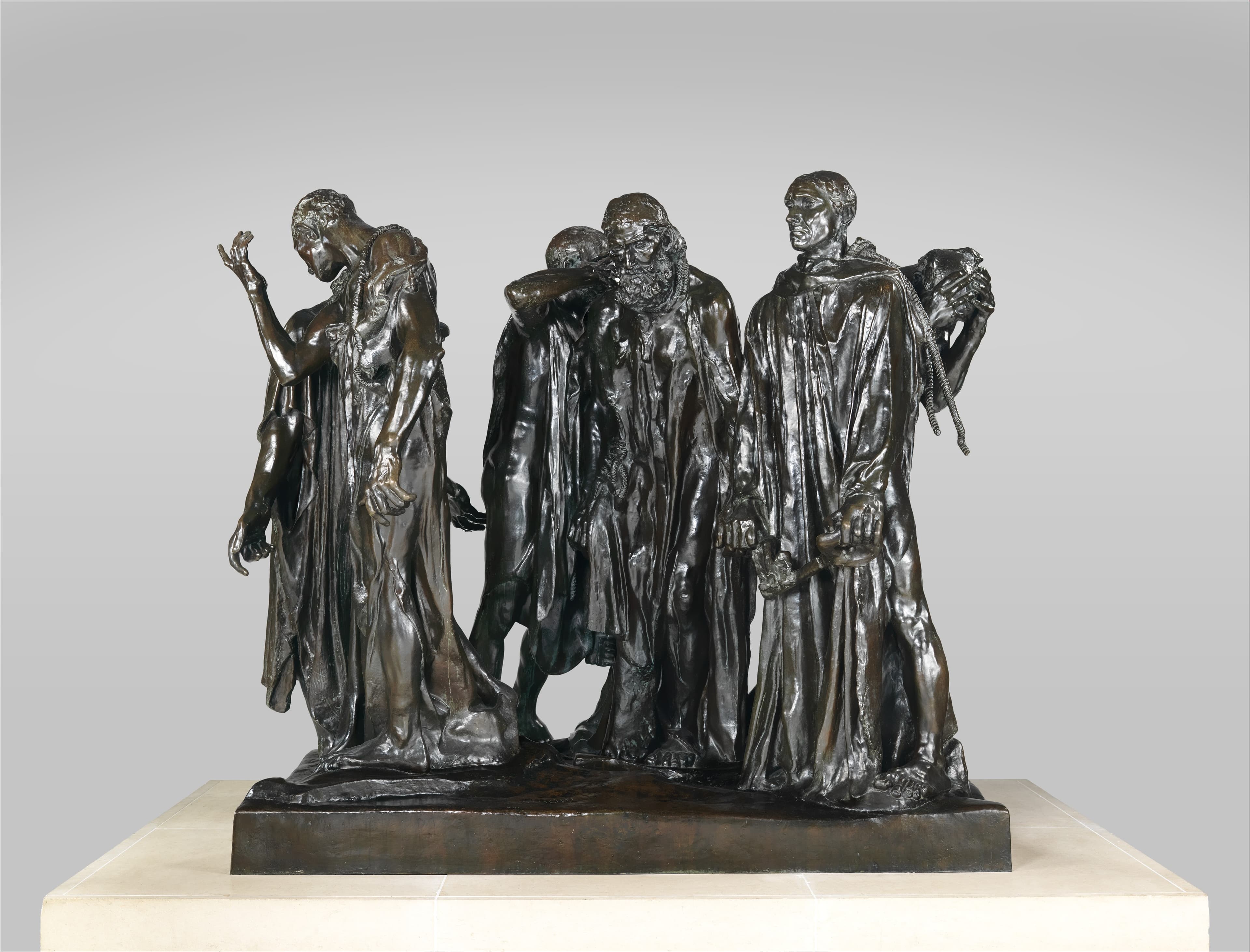 A bronze group of six men in long coarse robes standing close together with ropes around their necks. One at the left bows his head and raises an open hand with the fingers spread; a bearded man faces forward at the centre; a man at the right stands upright in profile with his jaw set; behind him a hand is clamped over a head. Two large hands hang low among the robes, one closed in a fist and one holding a large key. The patina is dark brown to black with silvery highlights on the folds