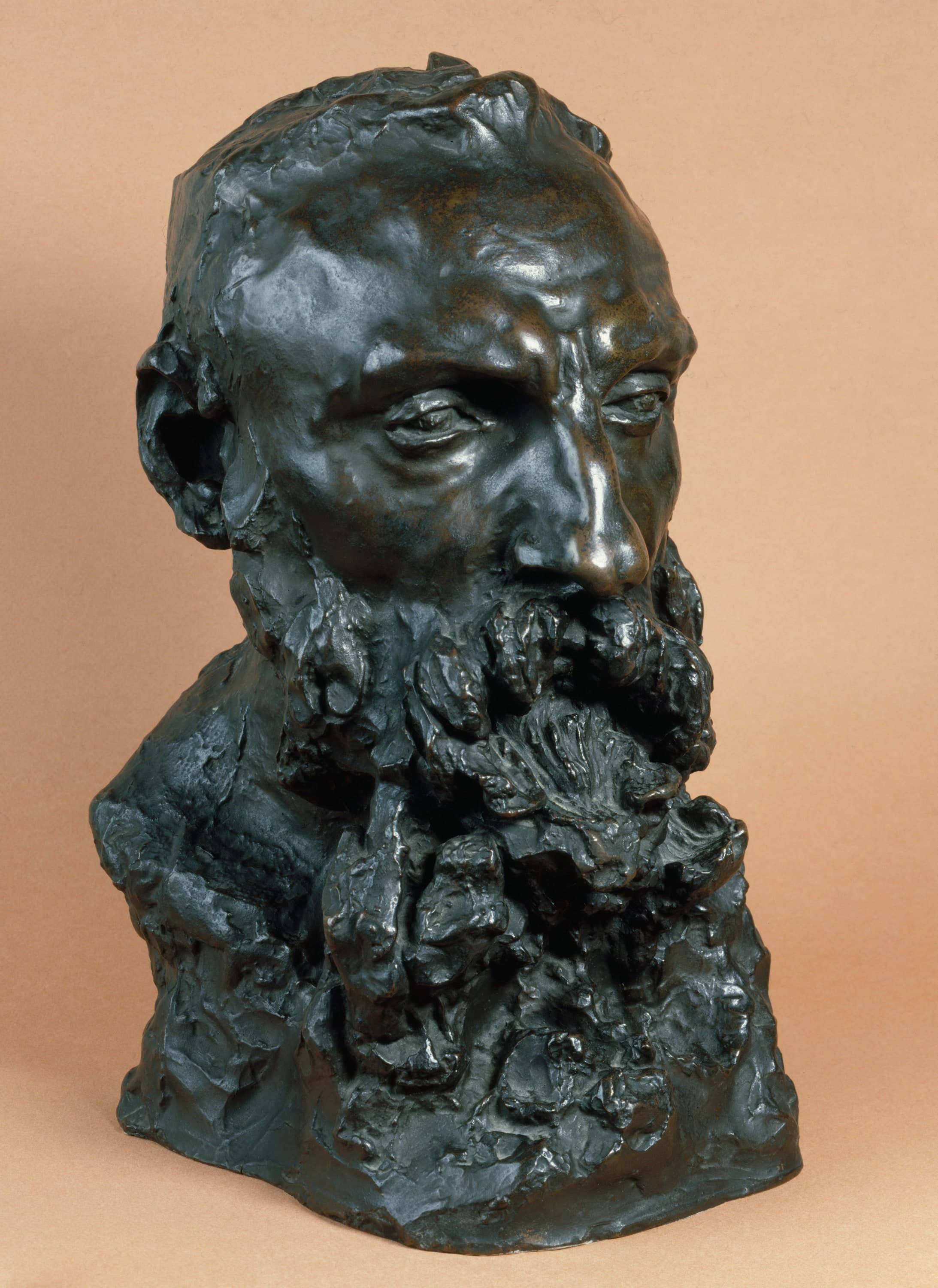 A bronze bust of a bearded man on a plain peach studio background, turned three-quarters to the viewer's right and lit from that side. The forehead is high and domed, the brows are heavy ridges and the eyes sit deep in shadow beneath them. The ridge of the strong nose carries the brightest highlight in the photograph. The beard is modelled as a mass of separate lumps of bronze that covers the whole chest. The left ear is a dark hollow. The bust ends in a rough torn edge below the bare shoulders, and the patina is dark green-black with brown and gold lights