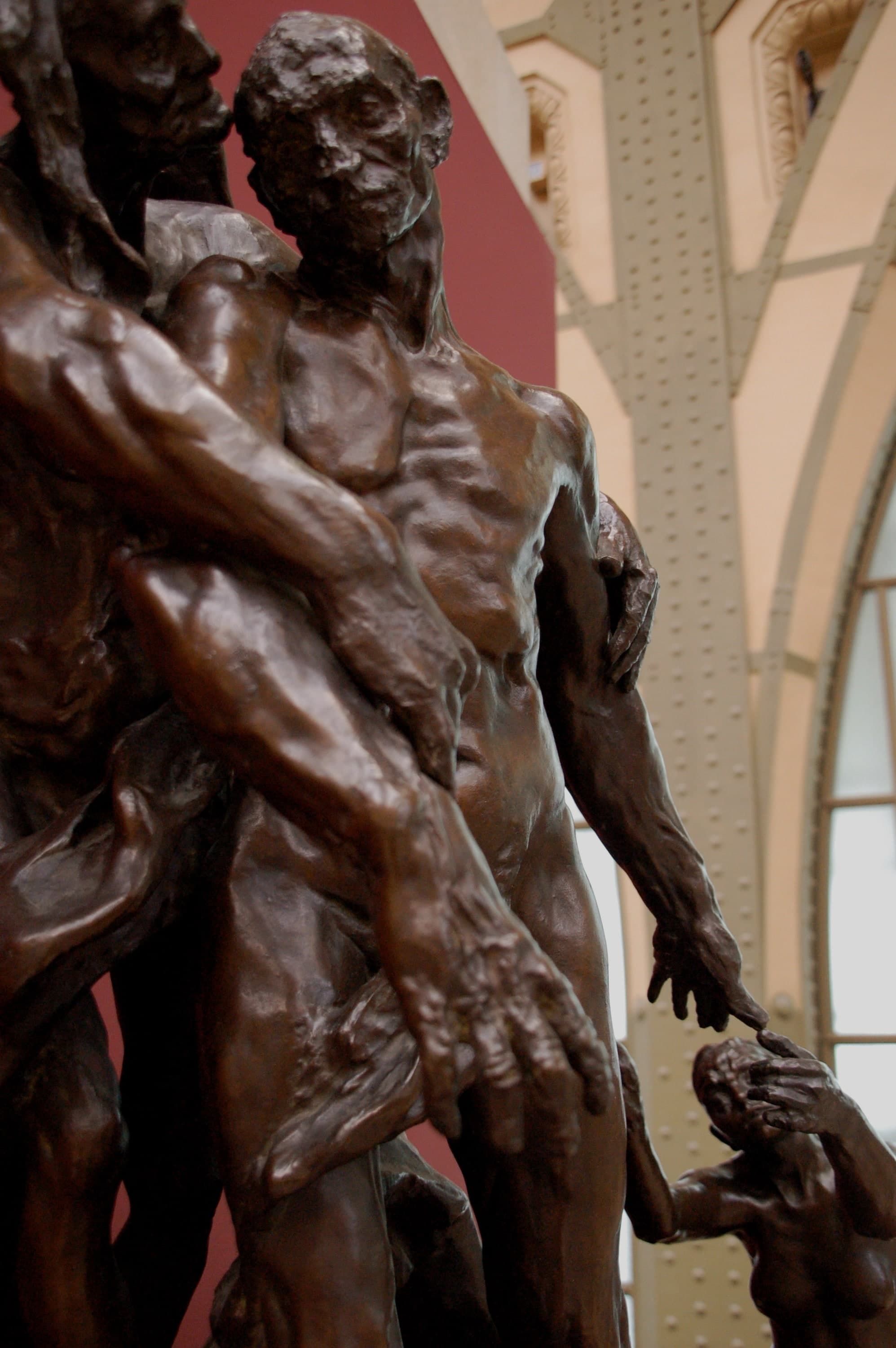 A low view of the bronze group in a museum gallery, filling the frame from the waist of the central man upwards. His head is bald and roughly modelled, tipped down and turned away to the left. Two thin arms reach in from the left across his chest and hip, the lower one ending in a large open hand with splayed fingers. His own arm hangs down at the right and its fingers stop just short of two small raised hands at the lower right corner. Cream-painted iron roof trusses and a dark pink wall stand behind