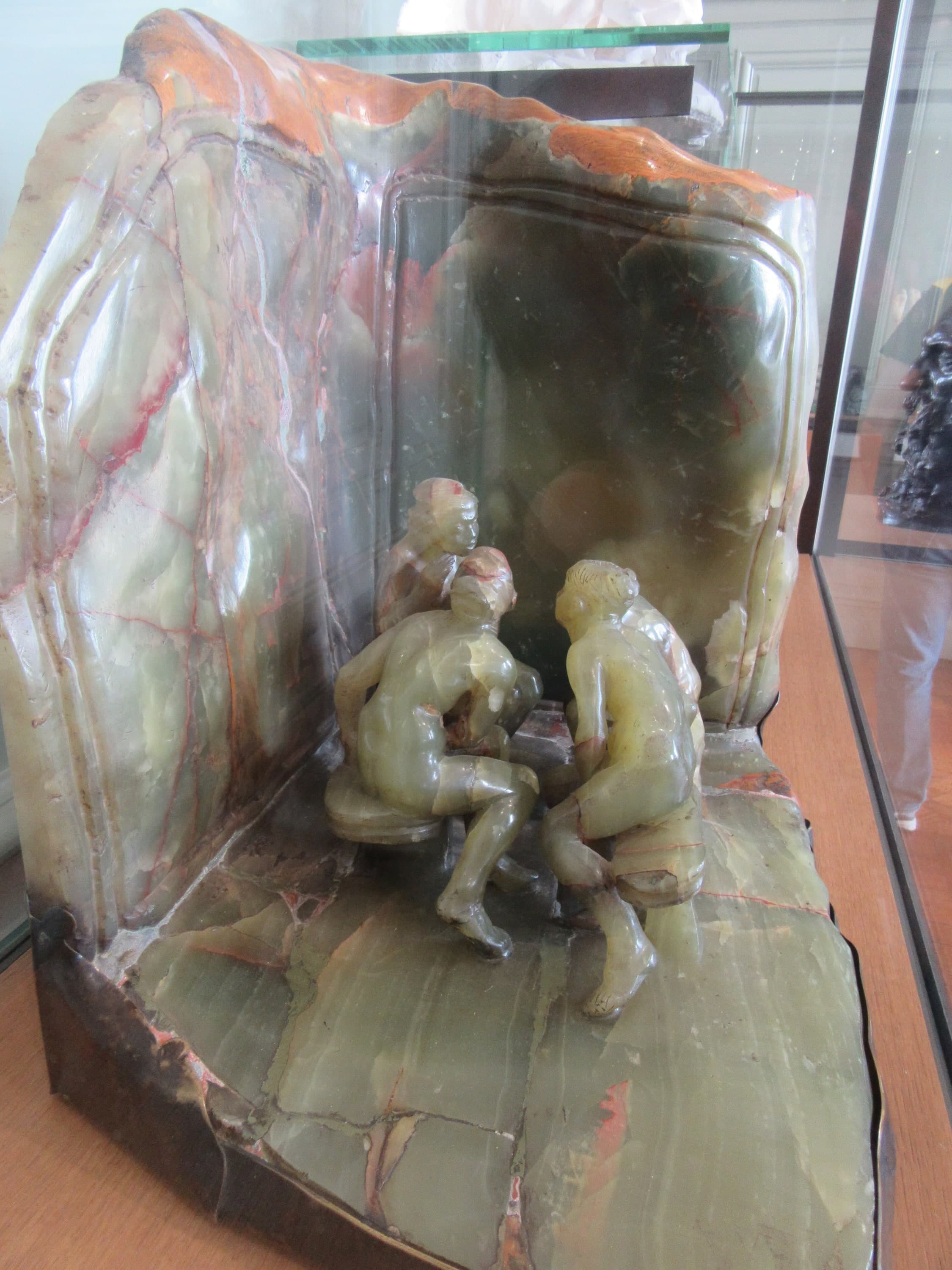 A small group carved in pale green onyx streaked with red and orange, photographed through the glass of a museum case standing on a wooden shelf. Two flat onyx panels meet at a right angle behind the figures, their top edge left rough and stained orange-red. Four small naked women are gathered in the corner: one sits on a low round stool with her back to the camera and her legs crossed in front of her, one leans in from the right with her head lowered, and two more are behind them. The floor of the group is a slab of the same veined stone, cracked in places, with a dark bronze edge along the front
