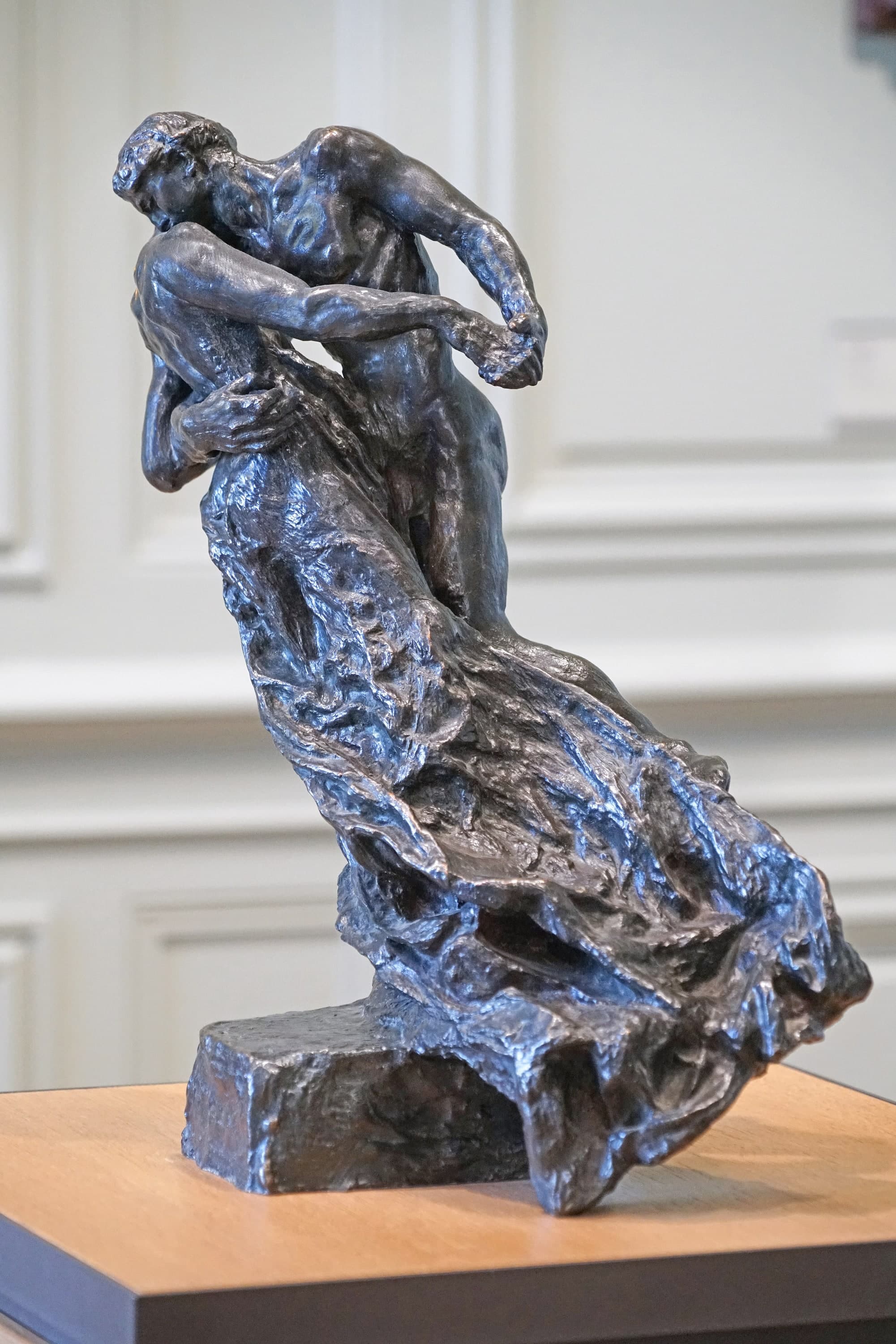 A small dark bronze of two dancers on a wooden museum tabletop against a pale grey panelled wall. The couple leans steeply to the left: the woman's head is tipped back against the man's shoulder, his head is bent over hers, one of his arms crosses her back and their hands clasp out to the right. A drapery starts at her hips and sweeps down to the right in deep folds, spreading into a wide hollow fan that reaches the end of the base. A plain rectangular block supports the left end