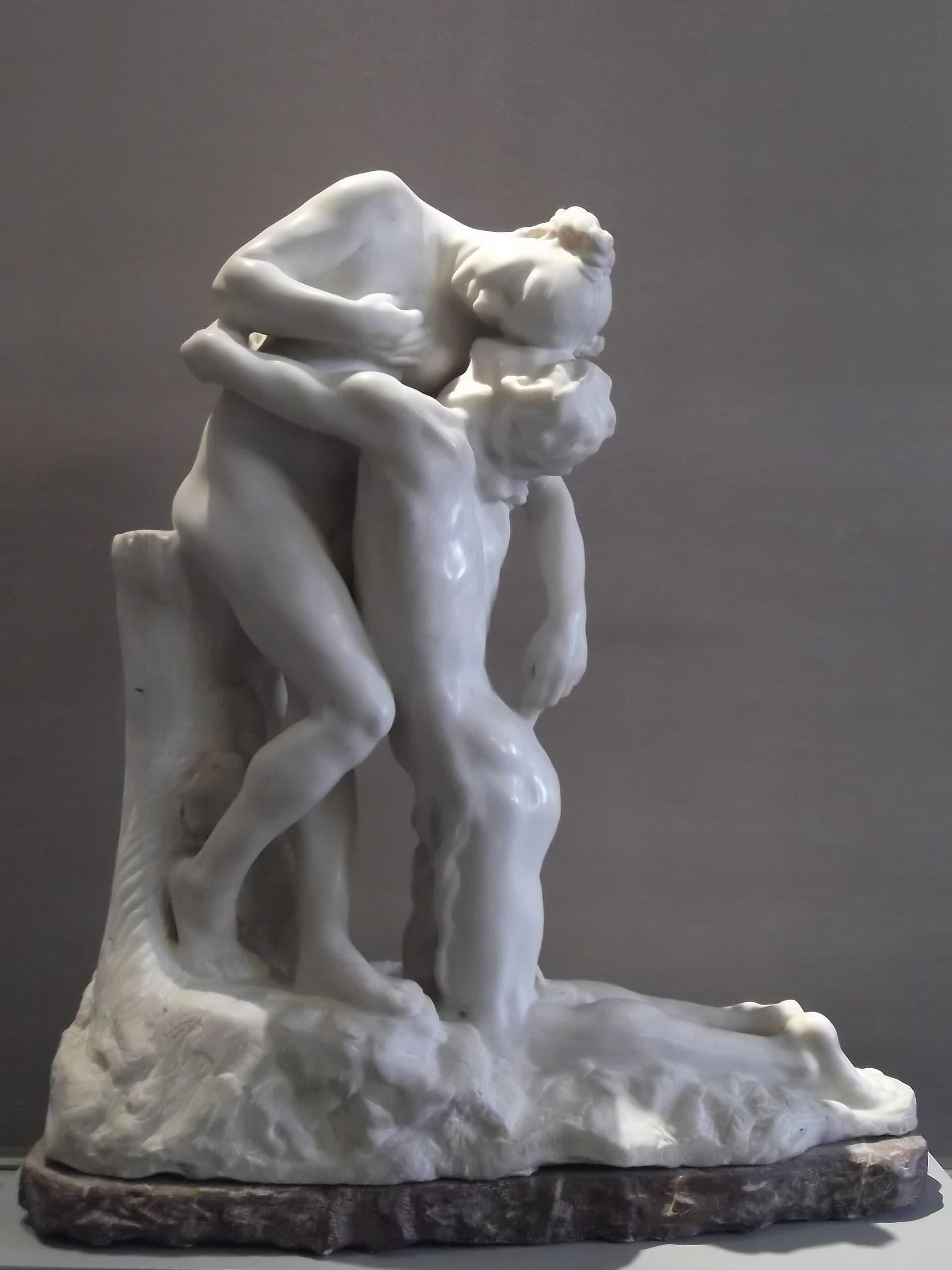 A white marble group on a rough white rock base set on a dark red-brown marble plinth, photographed against a plain grey wall. A young woman stands leaning back from the waist, her head tipped up and her eyes closed, one arm curved over the shoulders of a man who kneels in front of her with his face pressed to her chest and his arms round her hips. His legs trail back along the base to the right. A cut tree stump rises behind her at the left and supports her back