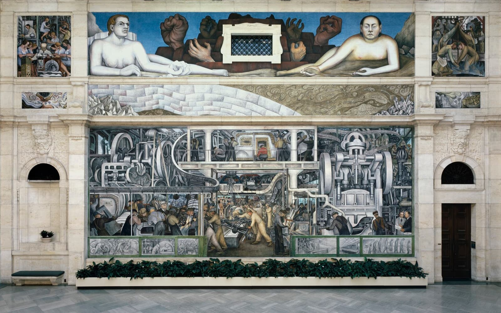 The south wall of Rivera Court photographed square on, the fresco set inside the museum's pale stone architecture with an arched doorway at the right and a planting box of foliage along its base. Along the top two nude figures recline facing inward, one pale-skinned with reddish-blond hair and one with East Asian features, with oversized brown hands pushing out of painted rock between them and a real stone window with a diamond-lattice grille at the centre. Below runs a band of pale limestone strata, white crystals and ochre fossil beds. The main panel is a body-production floor: a vast grey press with flywheels at the right, conveyors overhead, a worker in a red shirt seated in a car body on an upper gallery, crowds of workers in overalls, and two men in suits at a counter at the lower right. A strip of grey monochrome panels runs along the base behind a green rail