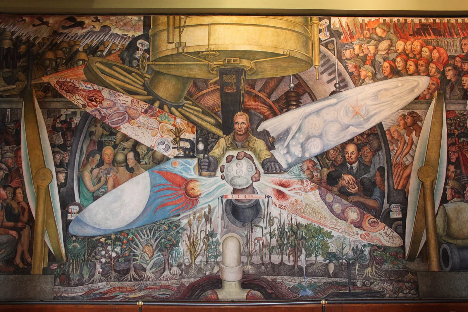 A mural photographed on the wall. At the centre a fair-haired worker in khaki overalls holds a lever in one gloved hand and a pale lensed sphere in both. Four long pointed ellipses cross behind him — one filled with cells and tissue, one with white spiral nebulae and a red star, one with an orange sun throwing red flares across blue, one with pale cellular forms. A brass drum of machinery hangs above him. At the upper left ranks of soldiers in gas masks advance with fixed bayonets under biplanes; at the upper right crowds in red headscarves sing under red banners before a crenellated wall and onion domes. Left of centre four women in evening dress sit at a card table with a bespectacled man behind them; right of centre a bald man with a reddish goatee in a dark suit joins the clasped hands of four workers. Along the base runs a band of crops with their roots drawn pale in striped earth