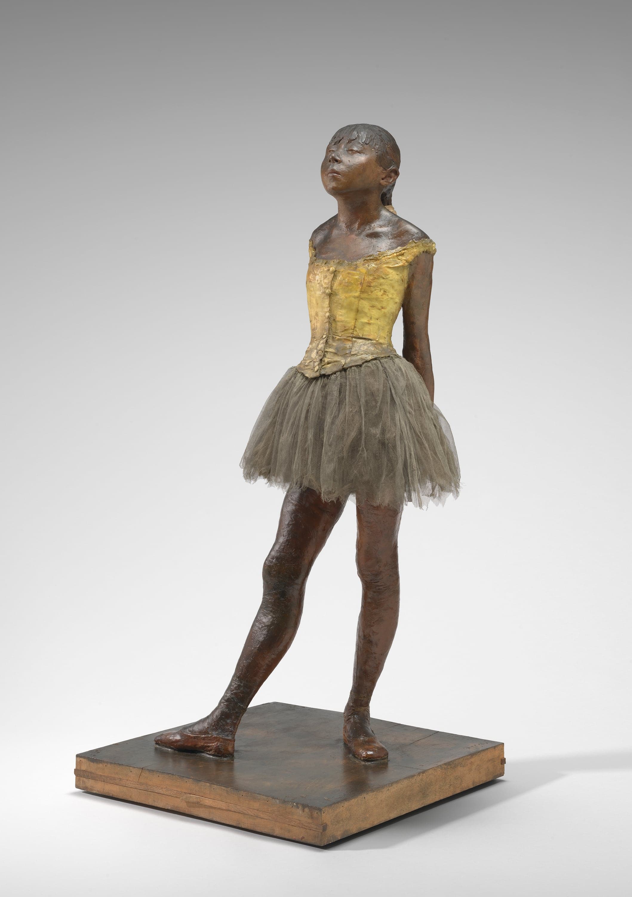 A wax statuette of a girl about two-thirds life size, standing on a wooden base against a gray ground. Her skin is a warm brown wax, her bodice is pale yellow, and she wears a real gauze tutu that has gone gray-green with age. Her hands are clasped behind her back, her chin is tilted up, her eyes are almost closed, and her right foot is turned out and set forward.