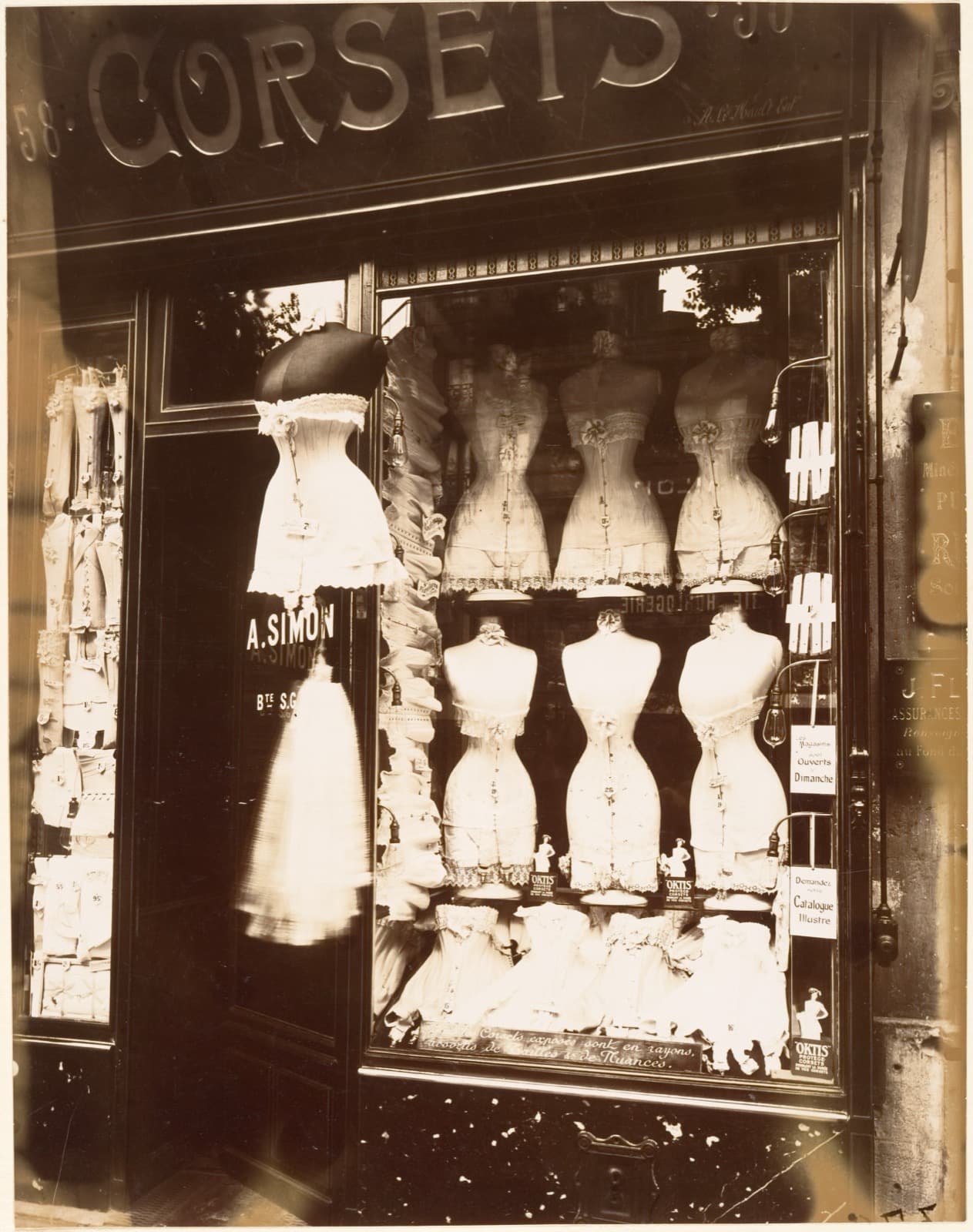 A shop window filled with headless dress forms wearing laced corsets, arranged in rows on stands and hanging from above, with price cards propped beside them
