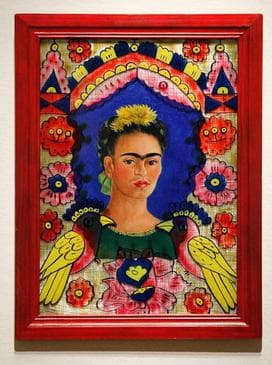 A small self-portrait photographed in its red-painted wooden frame against a pale wall. Frida Kahlo's head and shoulders face forward against a deep blue pointed arch, her eyebrows painted as one unbroken line, a cluster of yellow flowers pinned on top of her dark centre-parted hair, and a dark green blouse with gold trim at the neck. On the glass in front of the portrait a bird with a navy head, a yellow beak and one large yellow wing faces inward at each shoulder, a navy heart holding a yellow flower sits below the blouse, and stacked bands of navy, yellow, orange-red and dotted pink arch across the top