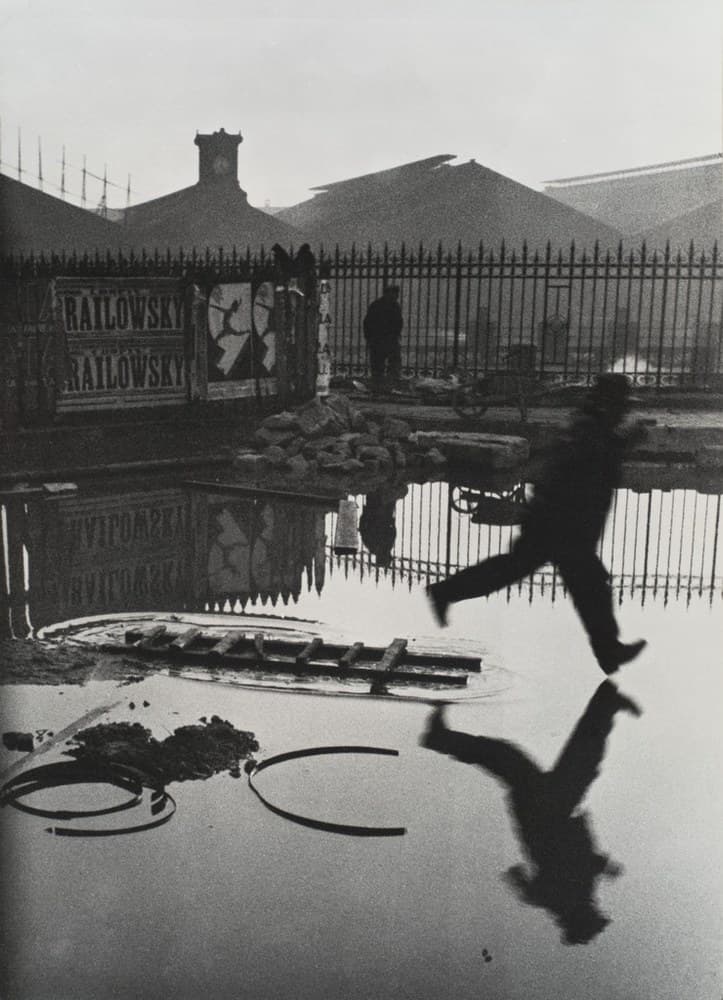 A man in a dark suit, dark shoes and a hat leaps from right to left across a flooded yard, his forward heel just above the water and his arms out, his legs and arms blurred by movement. His inverted reflection leaps beneath him. A wooden ladder lies half-submerged ahead of him inside an oval of ripple, and two metal hoops rest on the wet ground at the lower left. A tall spear-topped iron railing crosses the middle of the frame, with one man in a dark coat standing behind it beside a two-wheeled handcart and a heap of broken stone in front. A hoarding at the left carries the name RAILOWSKY printed twice in heavy capitals beside a torn poster of a leaping silhouette, both repeated backwards in the water. Above the railing rise three triangular train-shed roofs and a square clock tower under a flat pale sky